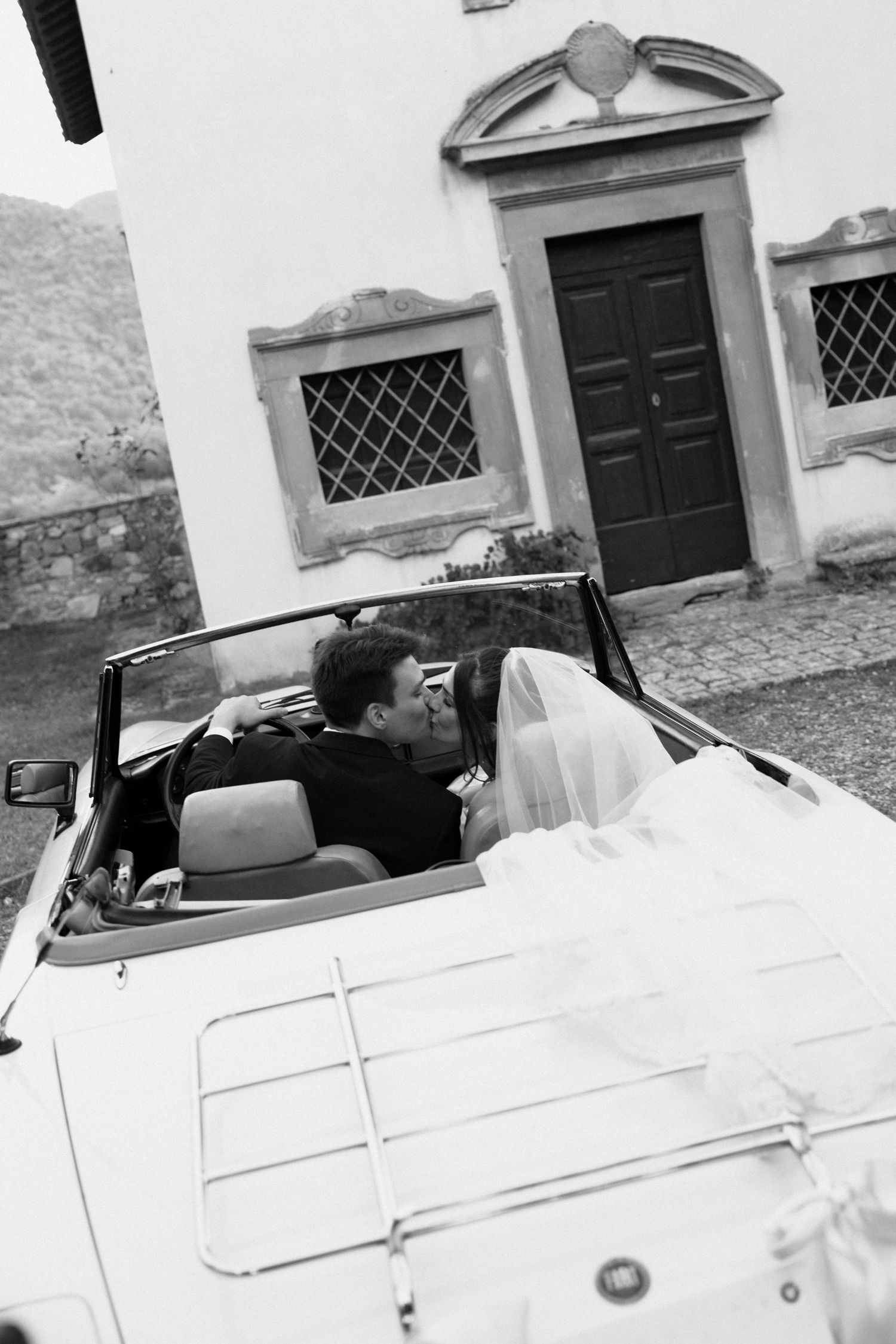 Black and white panoramic image of a classic white convertible car parked in front of a historic building facade.
