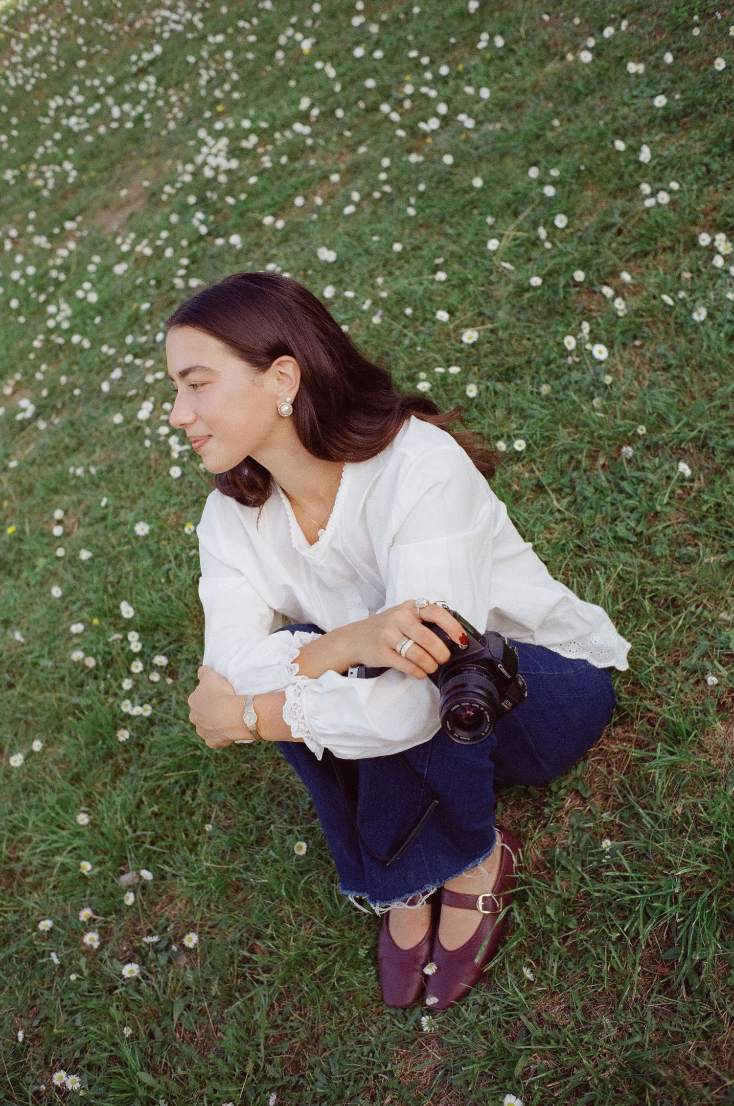 Person in white shirt and navy pants sits on grass with scattered white flowers in a candid outdoor photoshoot.