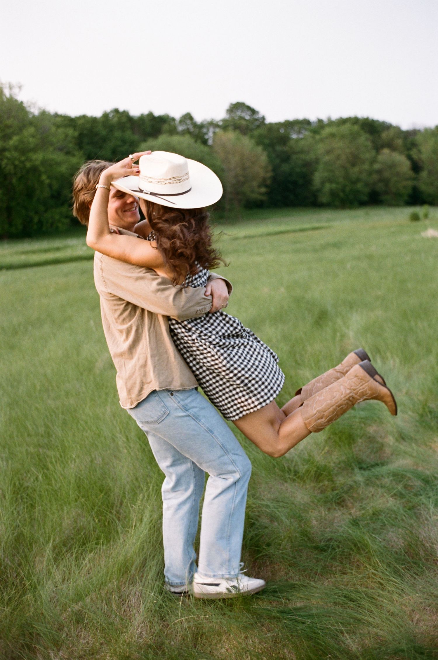 A couple embraces and spins playfully in a sunny green meadow wearing casual clothing and a cowboy hat.