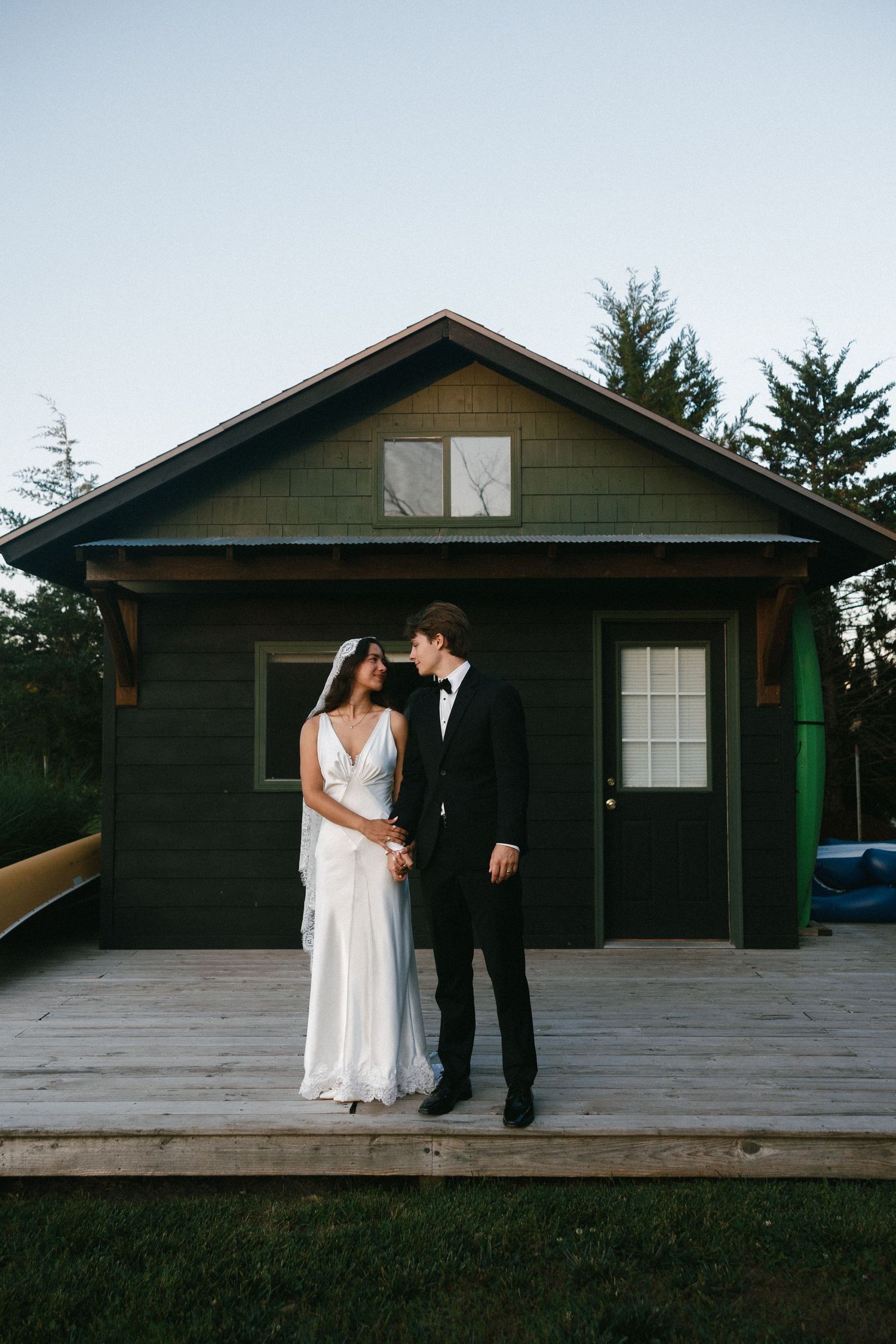 A rustic dark green cabin with a wooden deck and white trim set against a light sky.