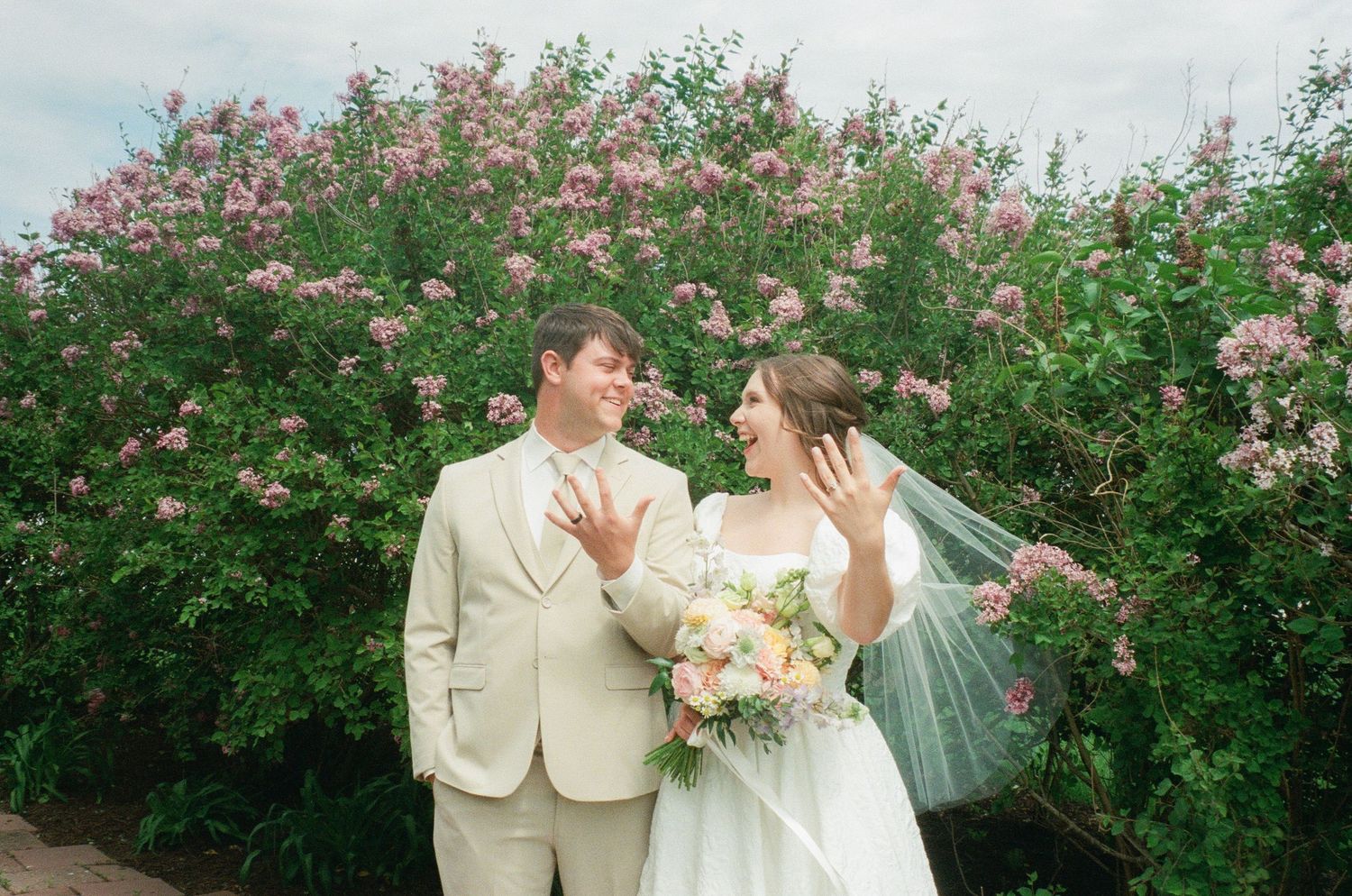 Newlywed couple showing off wedding rings in front of blooming pink flower bushes, bride holding pastel bouquet.