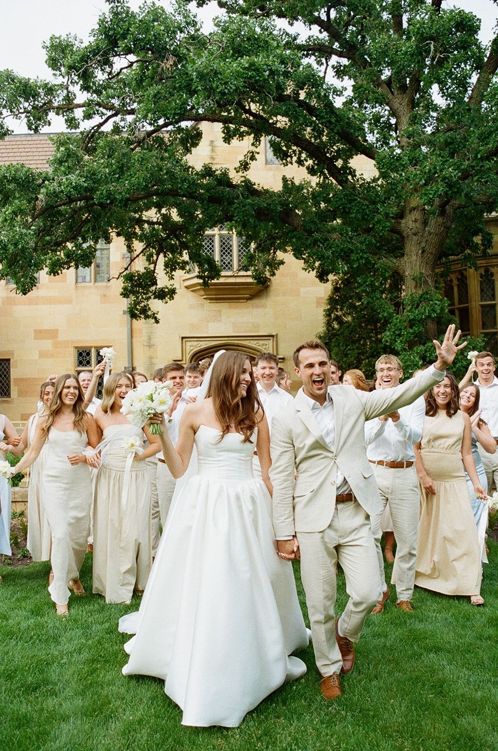 Wedding party wearing all white and beige outfits jumps joyfully on green lawn during outdoor celebration.