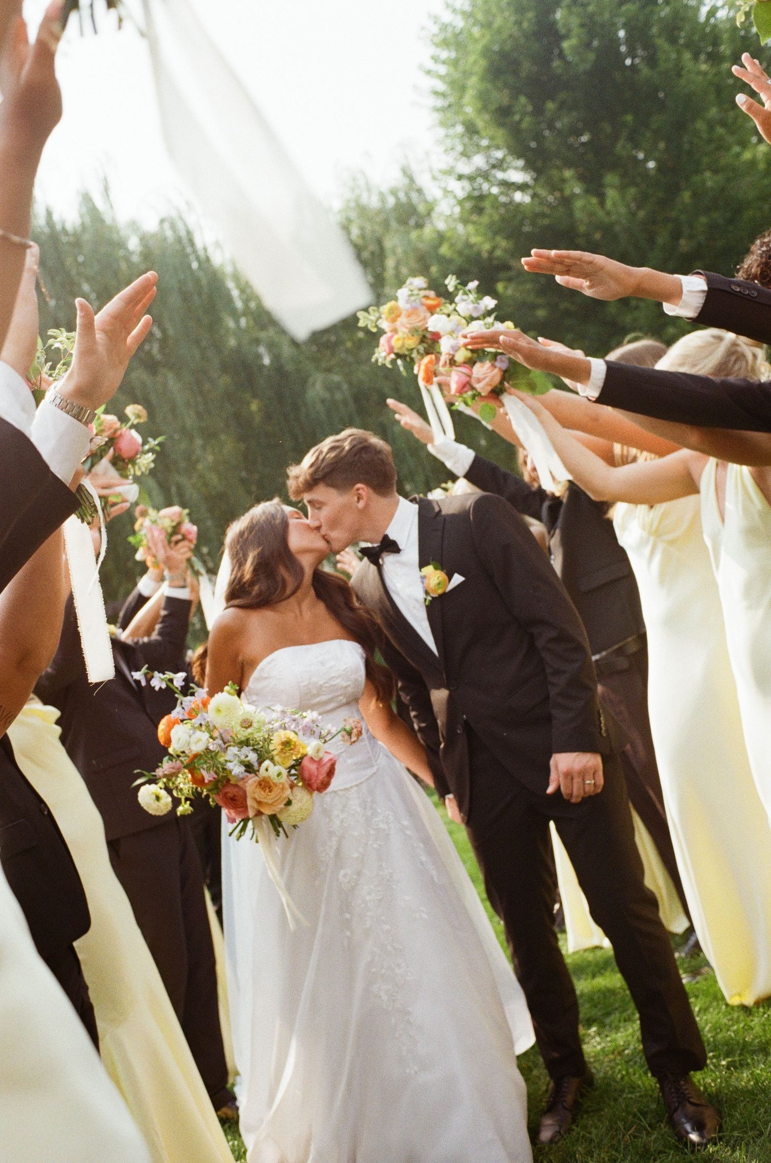 Wedding party in black suits and light dresses jumps joyfully together outdoors with flowers in hand.
