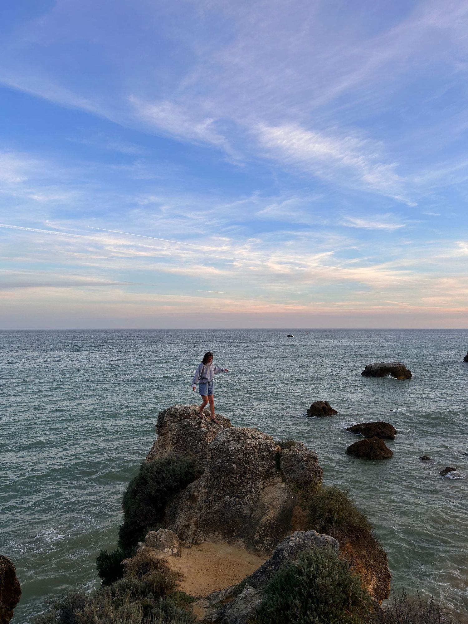 Person standing on coastal cliff overlooking ocean during sunset.