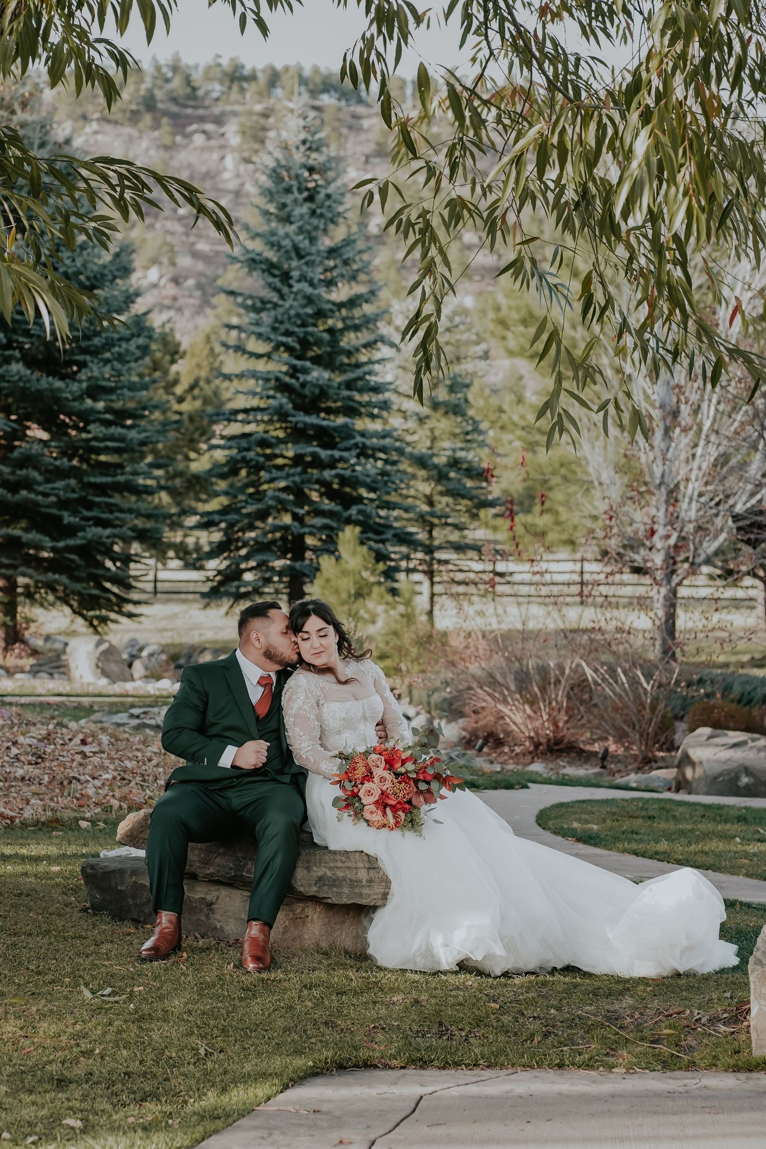 A romantic wedding couple sits together outside on a bench in a garden setting with evergreen trees and autumn foliage.