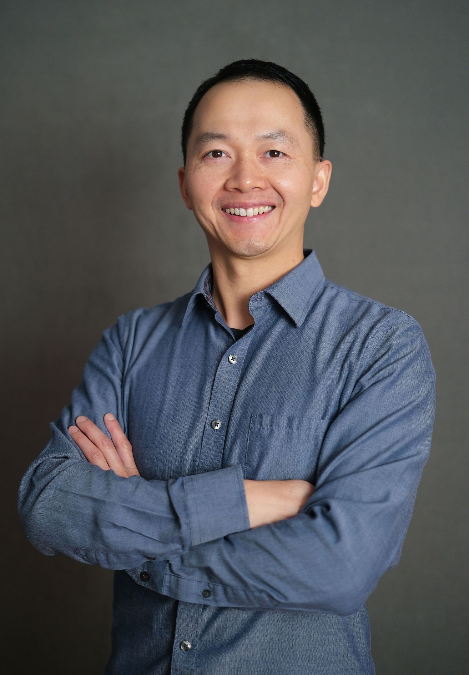 Headshot of a man wearing a blue shirt and crossing his arms and smiling, against a grey background