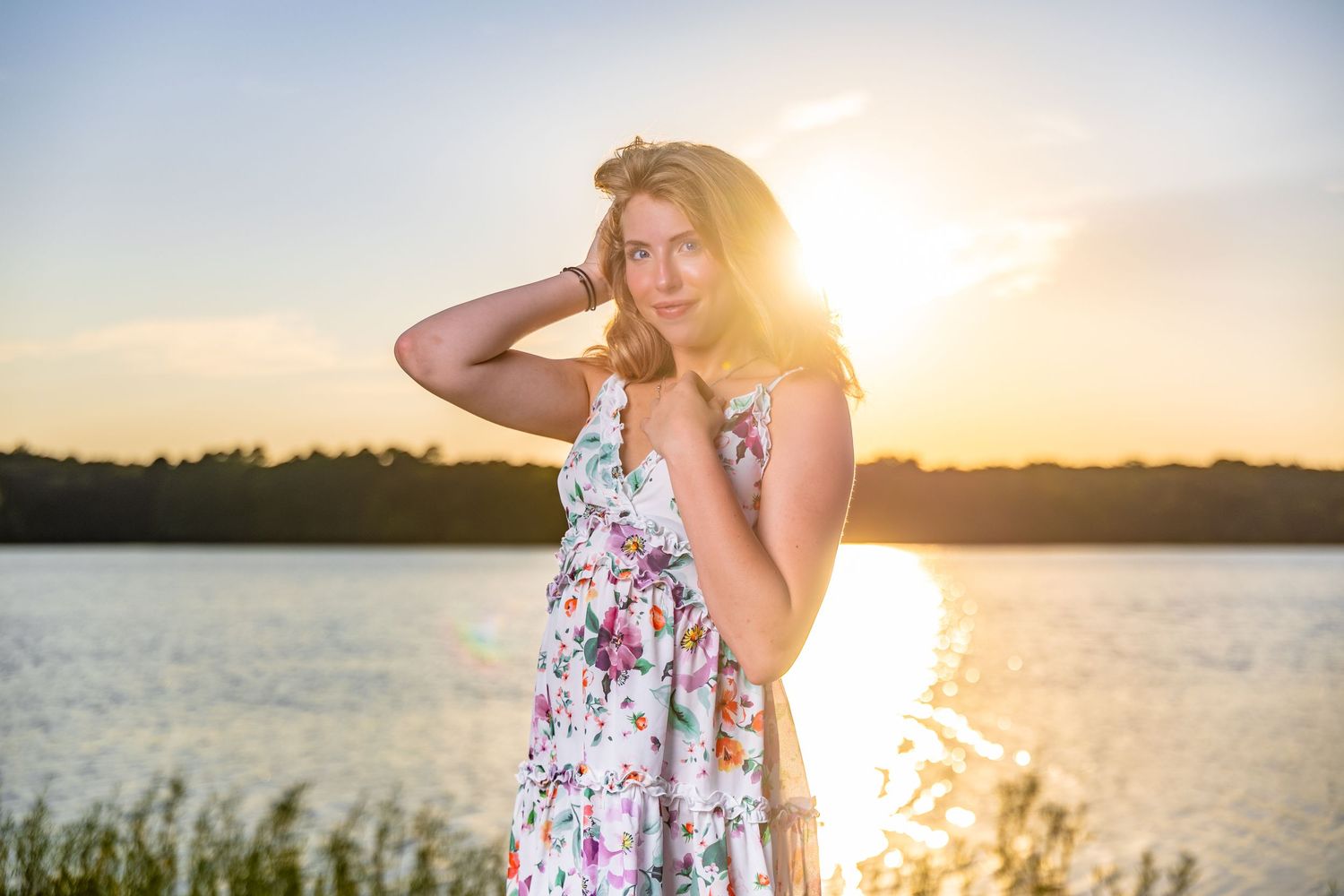 A woman in a floral sundress stands by a lake during golden hour sunset with sunlight creating a warm backlit glow.