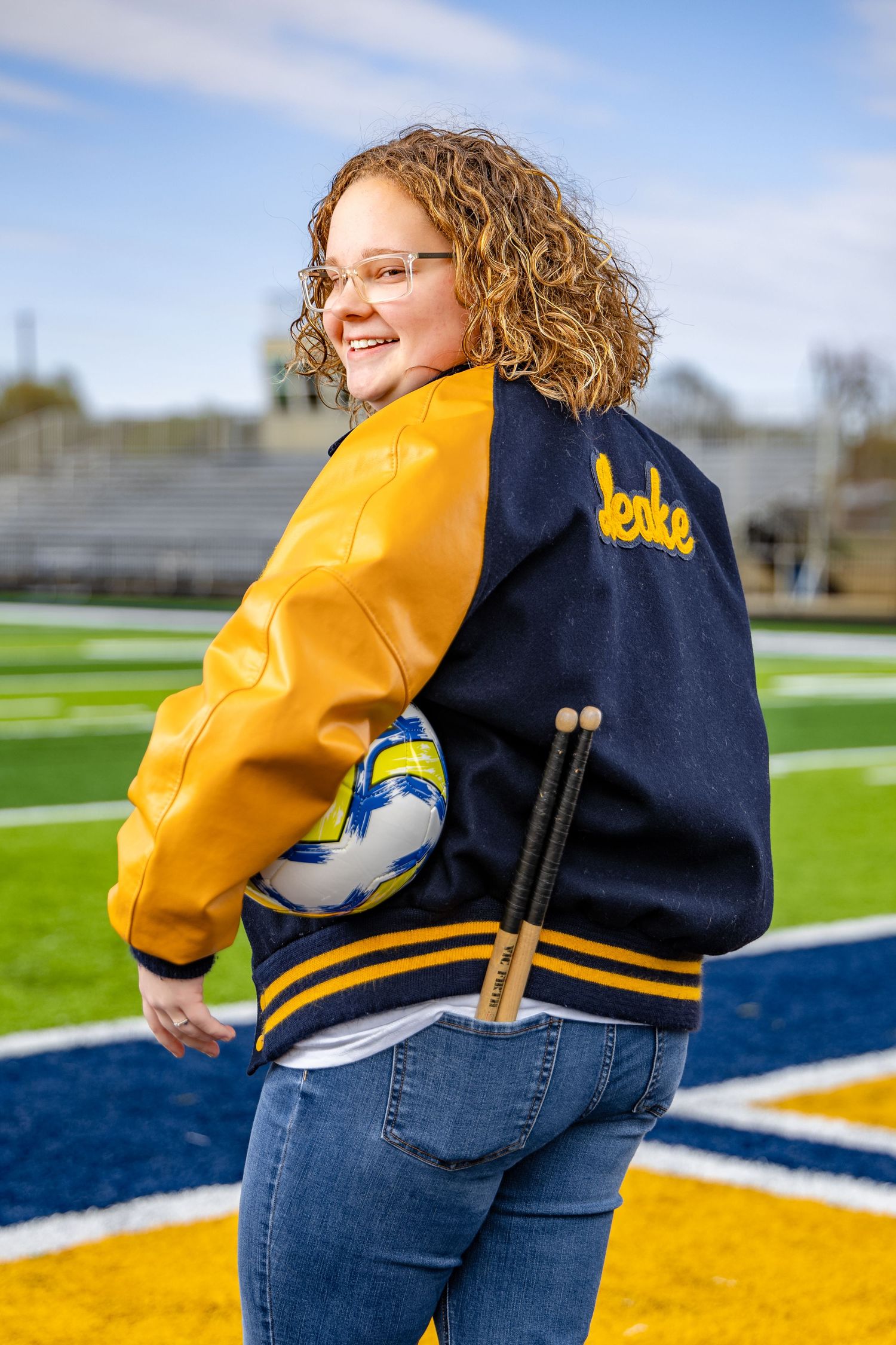 Athletic letterman jacket in navy and gold shown on a football field with blue and yellow markings.