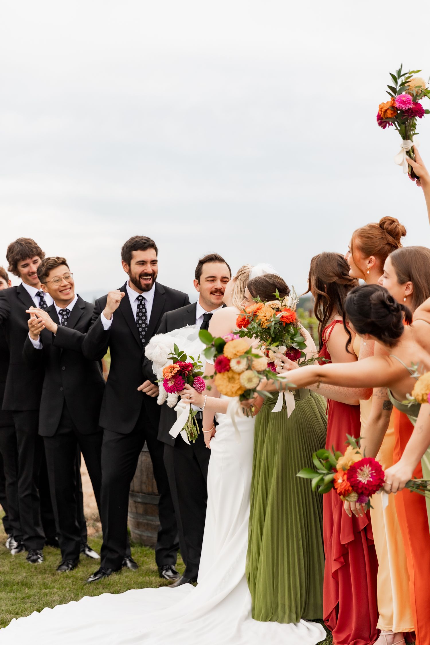Wedding party in colorful attire celebrates joyfully outdoors with bridesmaids in green and orange dresses.