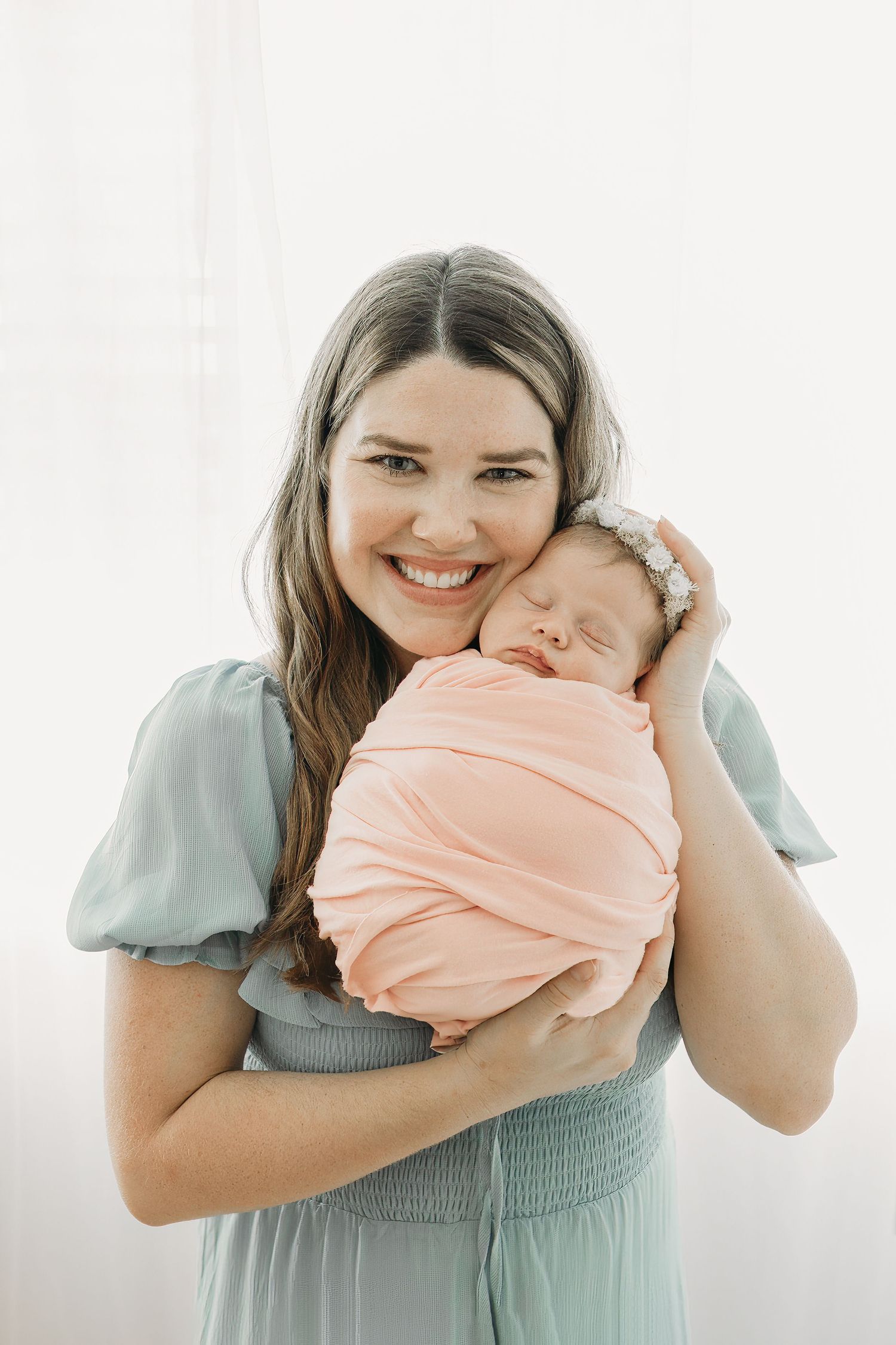 A new mother in a mint green dress holds her sleeping newborn baby wrapped in a peach swaddle against a white background.