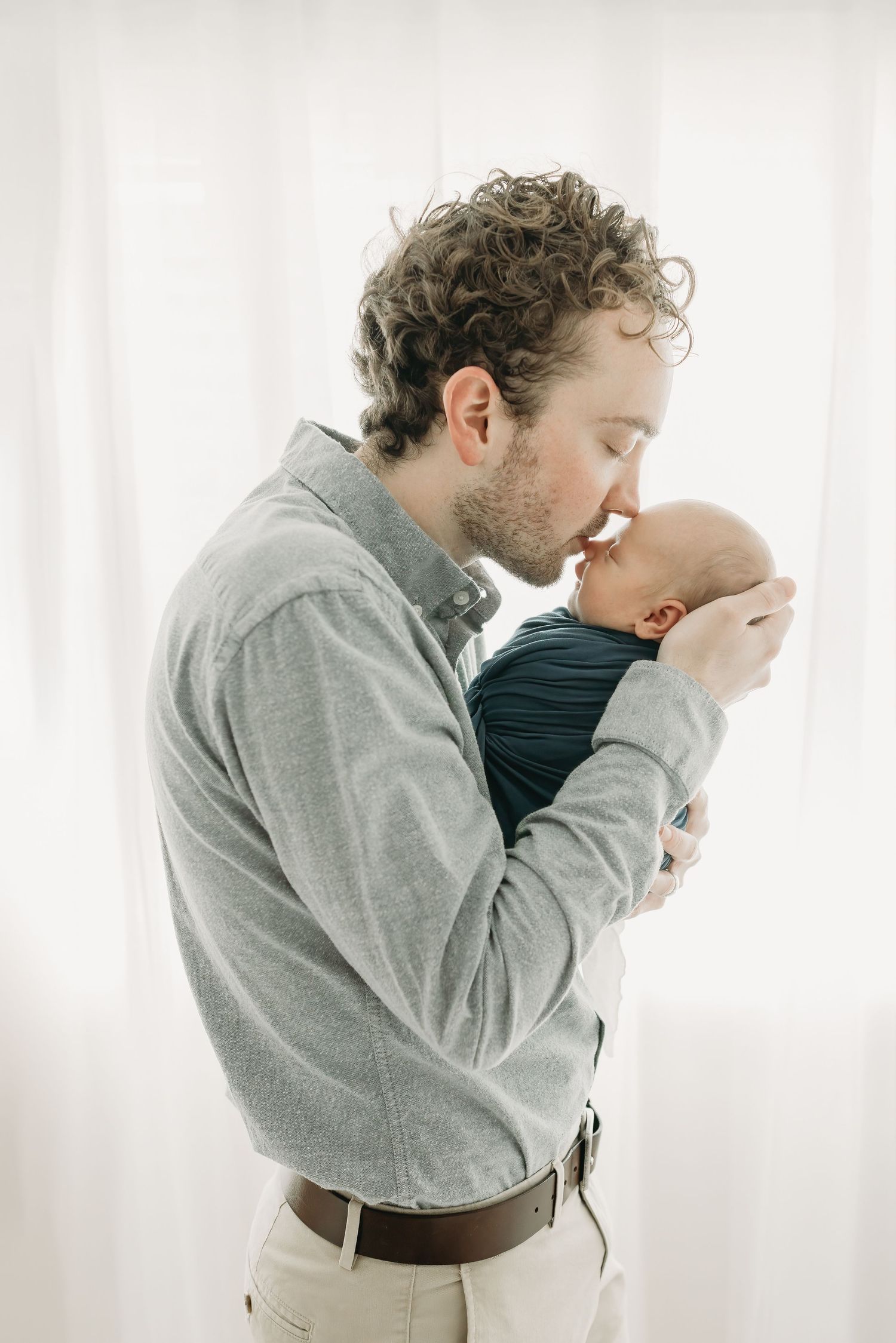 A tender moment between an adult in a gray button-down shirt holding and kissing a small infant wrapped in navy fabric.