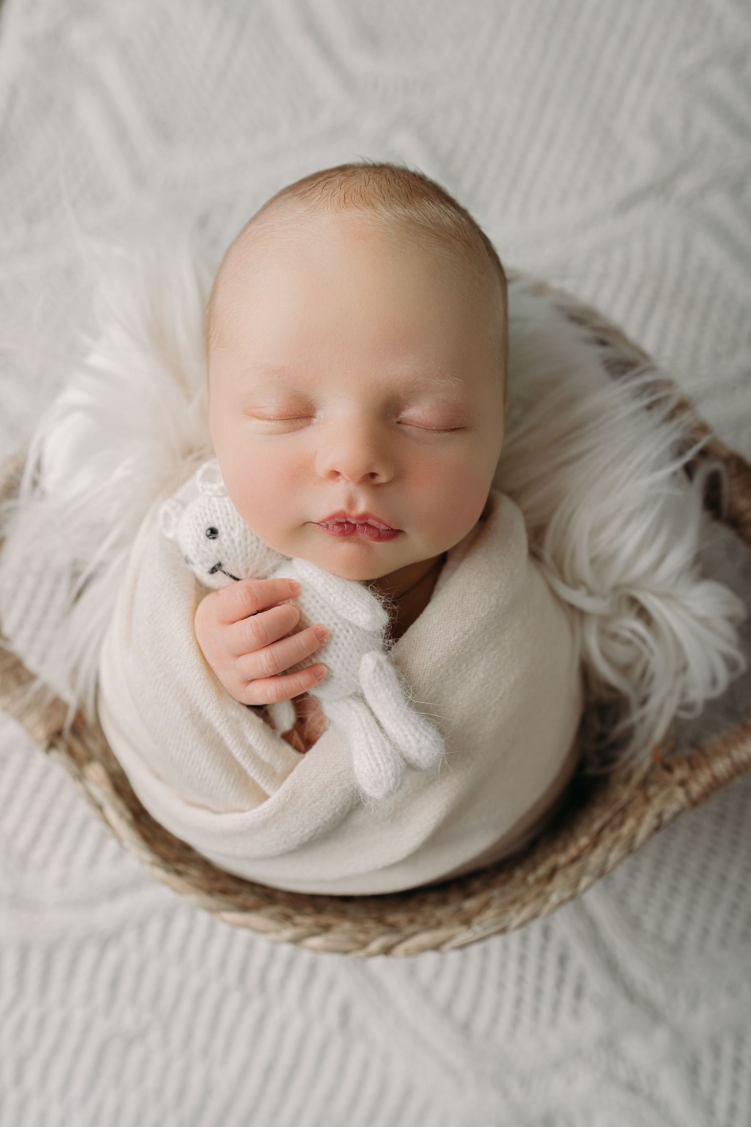 A sleeping newborn in white attire lies peacefully on round woven mat with plush toy in soft studio lighting.