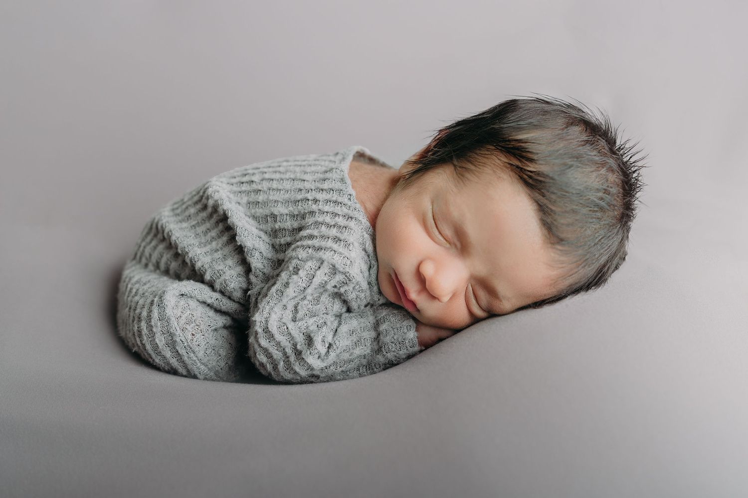 A sleeping newborn wearing a cozy grey knit sweater lies curled up on a grey backdrop during a professional photo session.