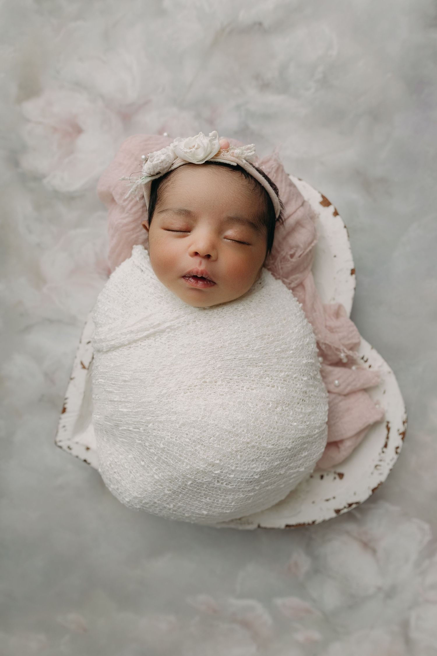 Sleeping newborn swaddled in white fabric lying on soft pink and gray background in portrait style.