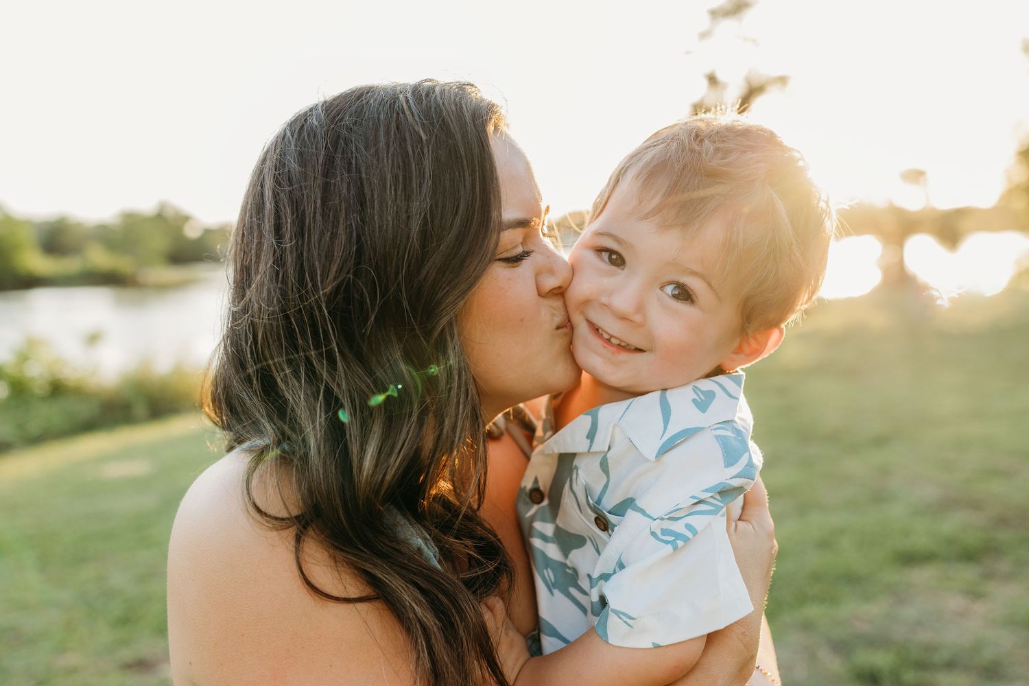 A tender moment between parent and child is captured during golden hour in a grassy outdoor setting by a lake.