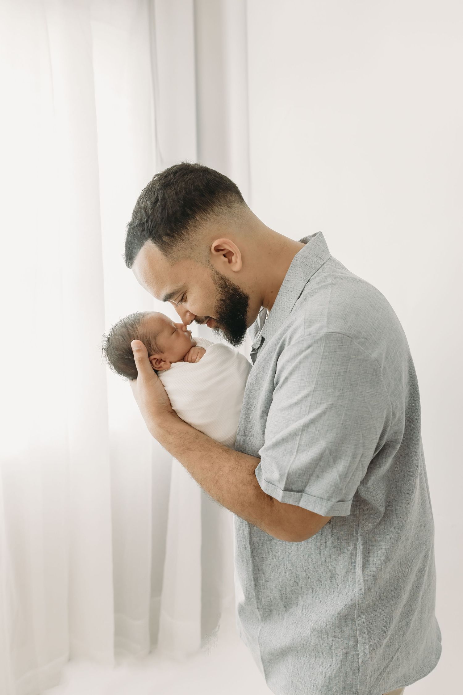 Father in grey shirt tenderly kisses newborn baby boy wrapped in white blanket during Tampa Newborn photos