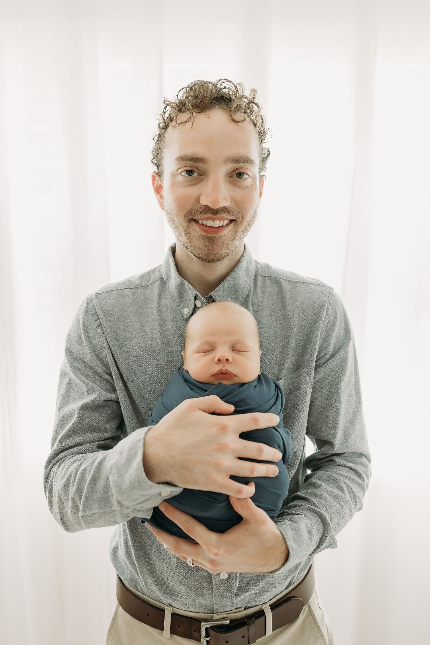 A parent in a grey button-down shirt tenderly holds a sleeping newborn baby wearing navy blue clothing against a white background.