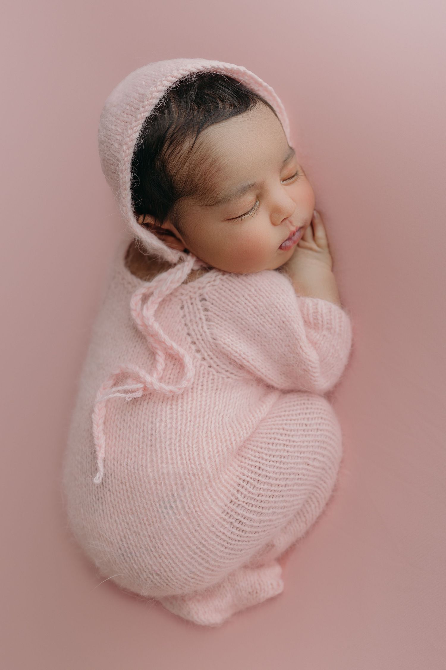 Sleeping newborn wearing soft pink knit bonnet and sweater curled up on pink background during professional photo shoot.