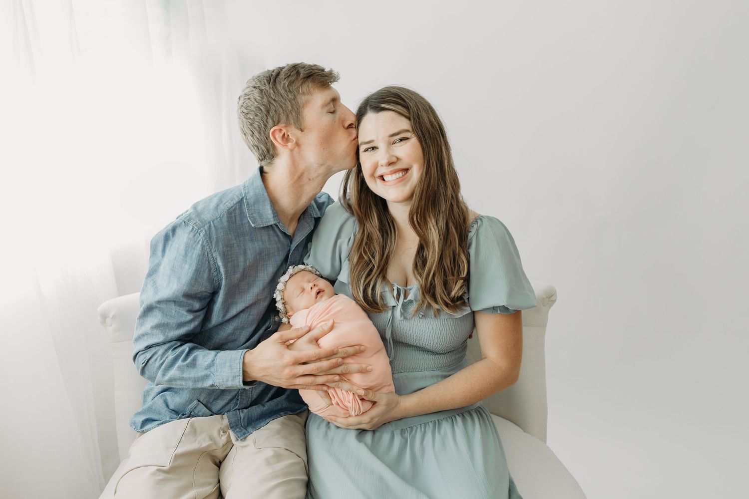 A couple in coordinating blue outfits snuggle together on a white background while holding their newborn baby.