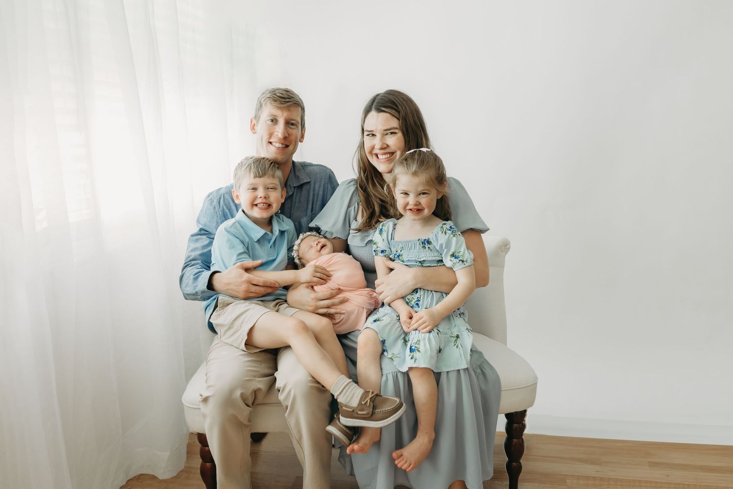 A family dressed in coordinating blue and white outfits sits together on a white settee against a light background.