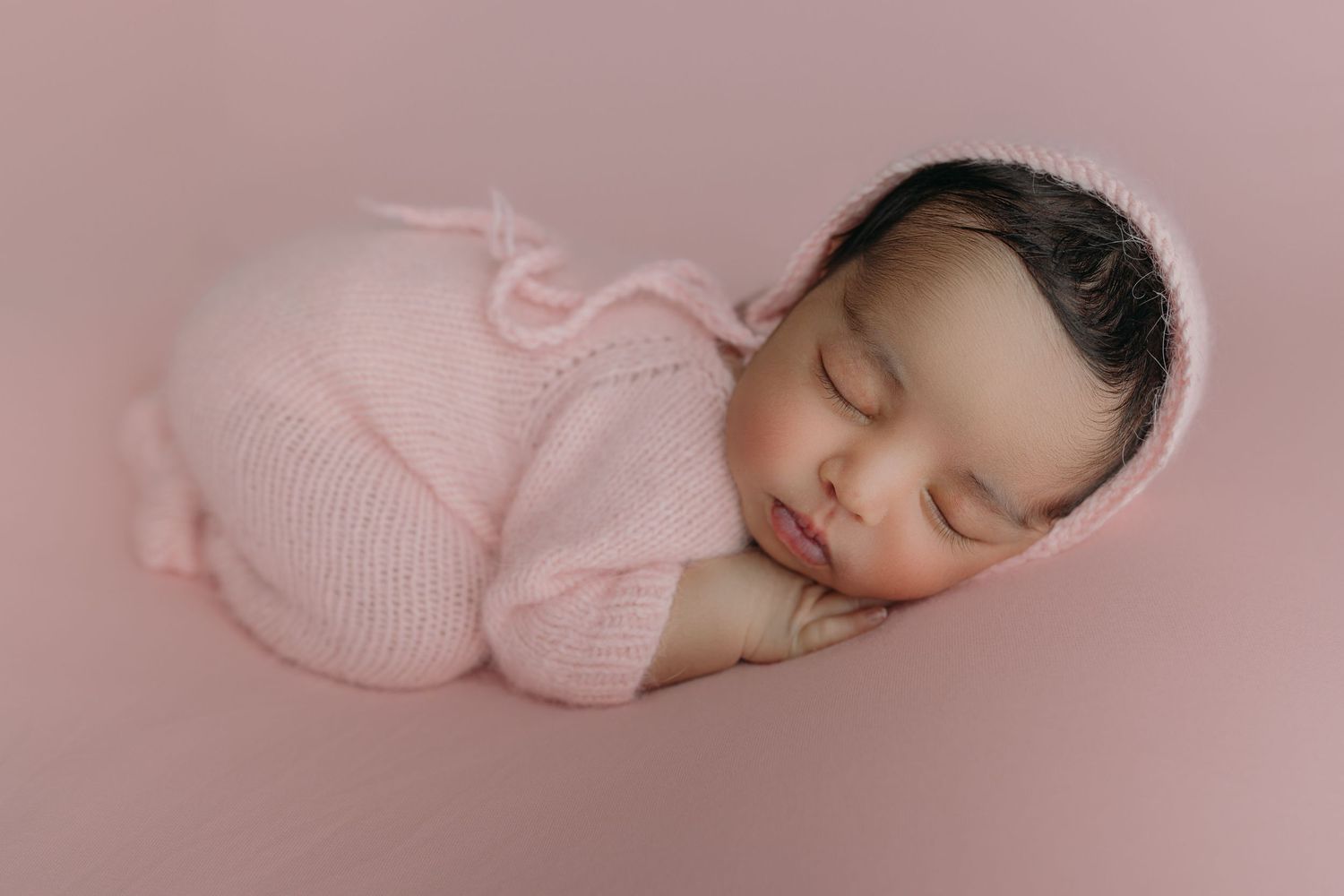Peaceful sleeping newborn baby wrapped in soft pink knitted romper on pink background.