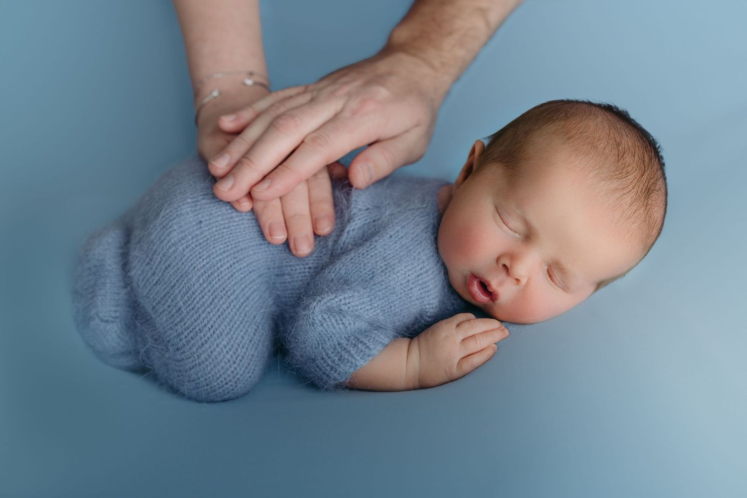 Sleeping newborn in blue knit outfit on light blue backdrop with hands gently touching their back.
