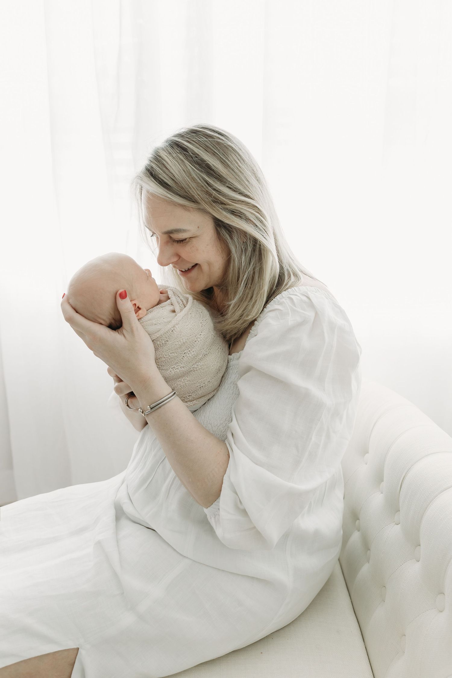 A tender moment between mother and newborn baby in white clothing sitting on a tufted couch by a window.