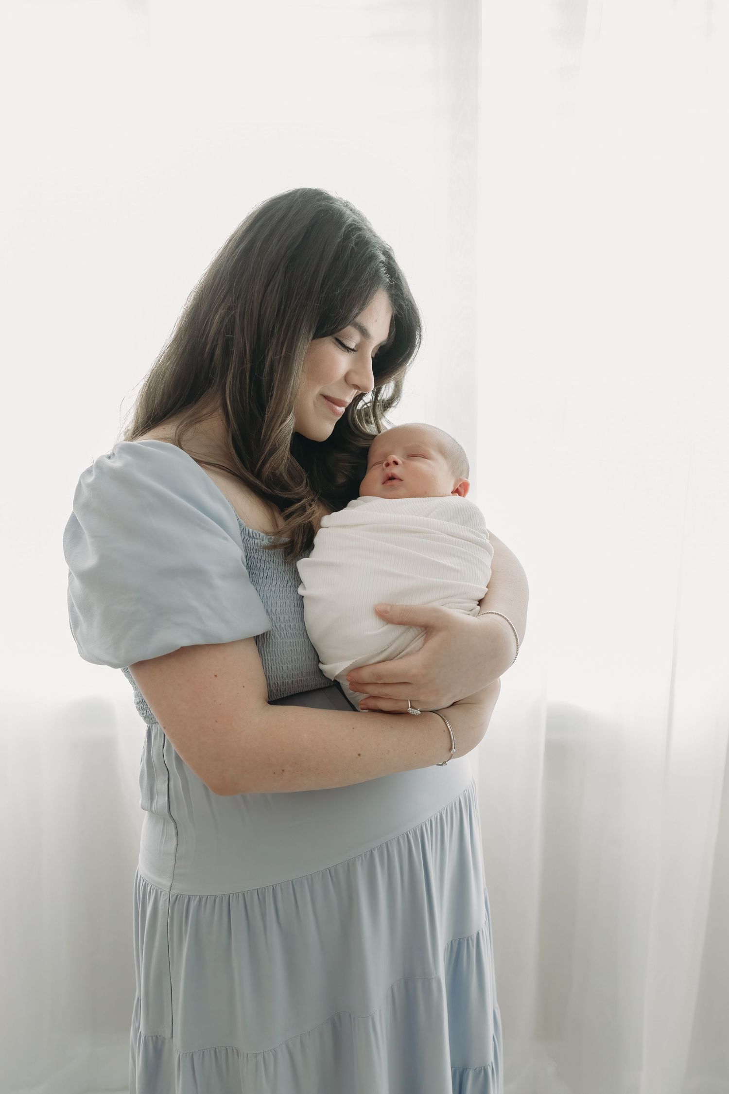 Mother in light blue dress tenderly holds and cuddles newborn baby in white swaddle against bright window backdrop.