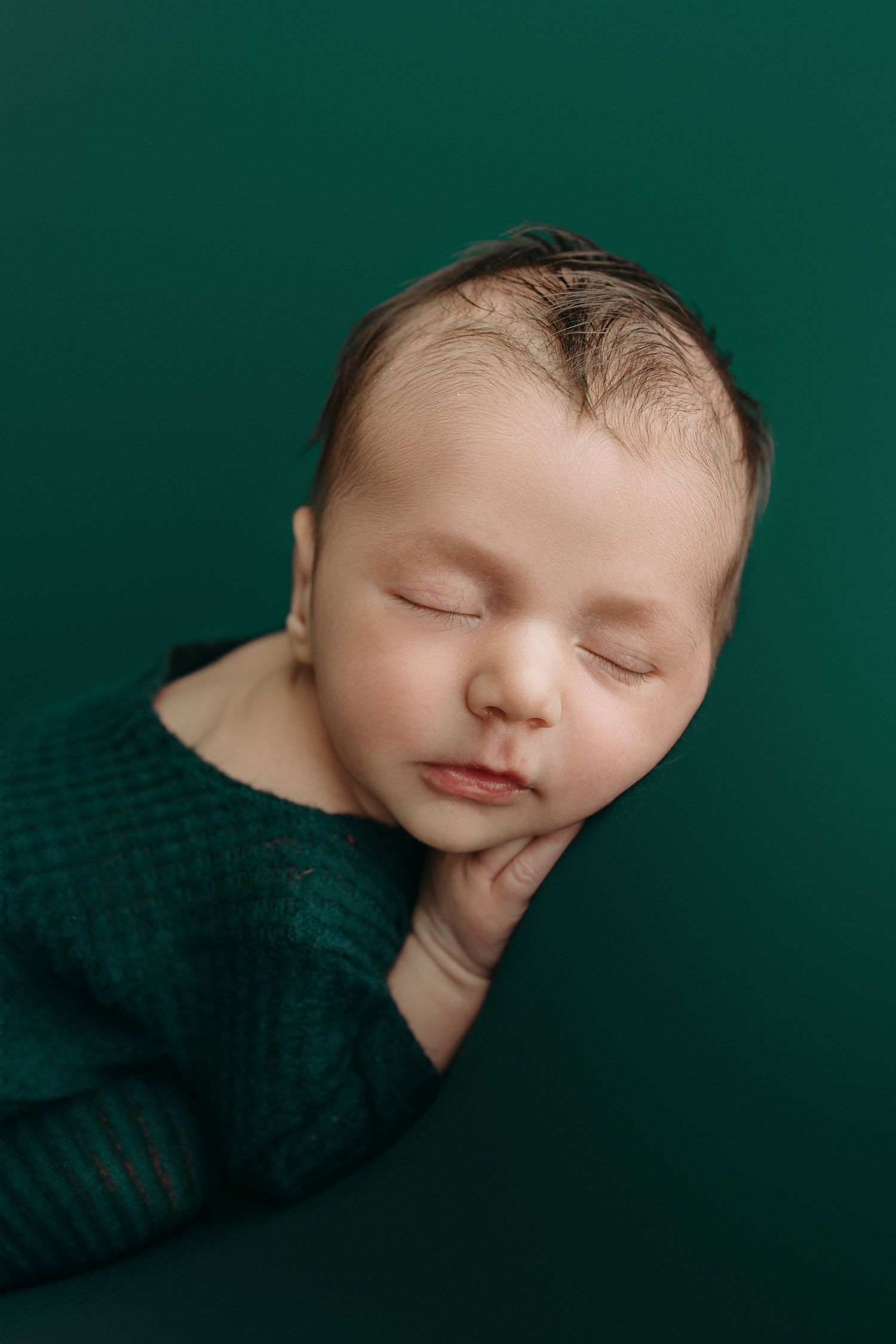 Sleeping newborn in dark emerald green wrap against matching background, with soft lighting and peaceful expression.
