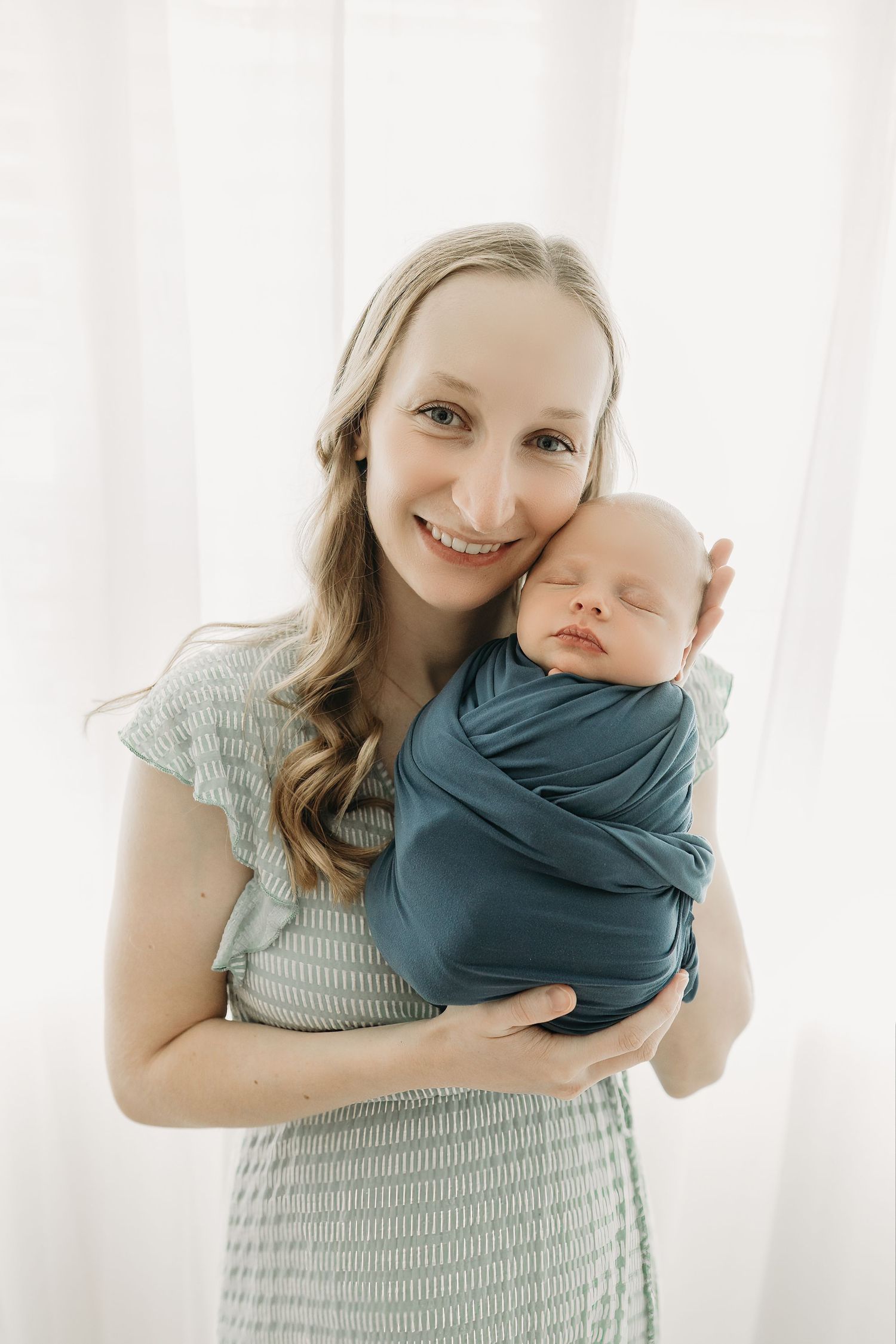 A loving parent holds their sleeping newborn baby wrapped in teal swaddling against a light background.