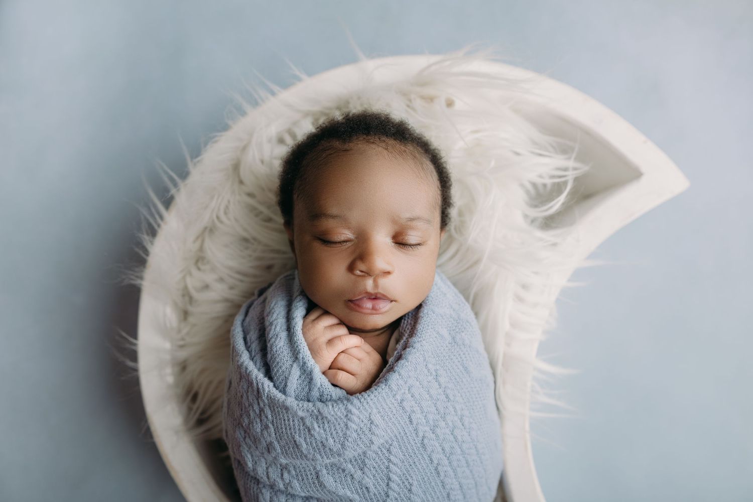 Sleeping baby swaddled in a blue knit blanket rests peacefully in a white circular cushion.