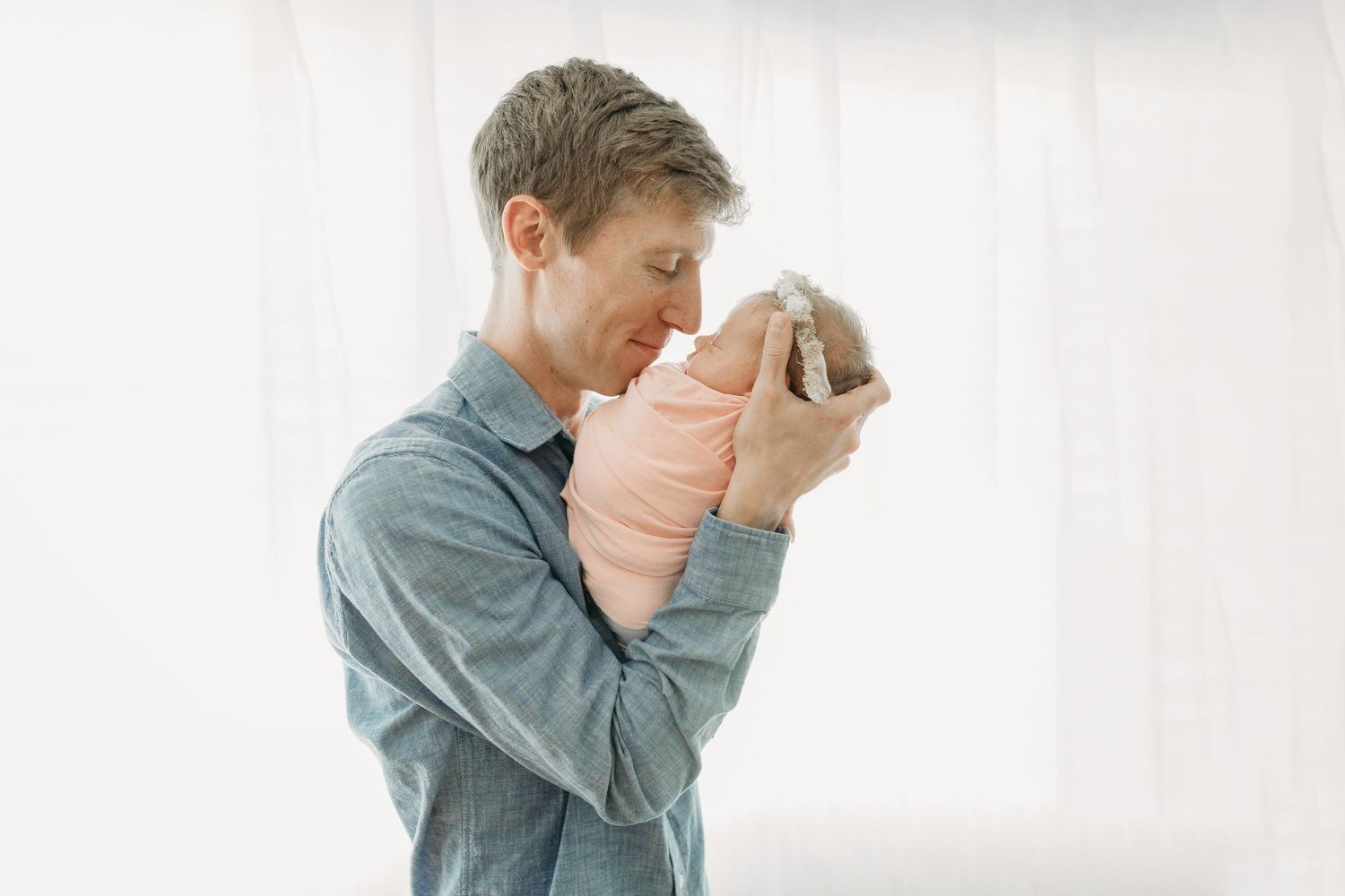 A person in a chambray shirt tenderly holds a newborn baby wrapped in a light pink blanket against a bright window.