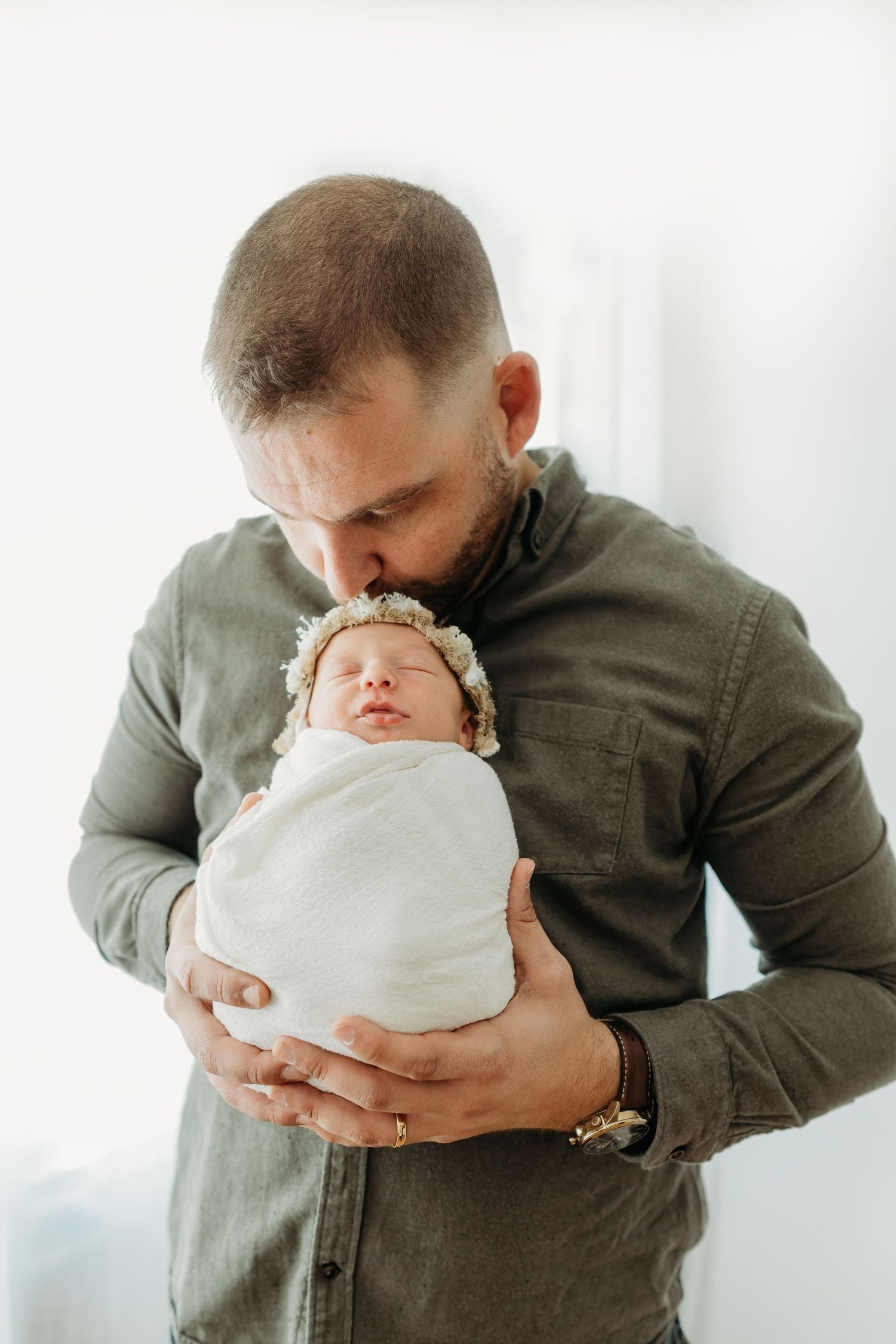 A parent in an olive button-down shirt tenderly holds a sleeping newborn swaddled in white against a light background.