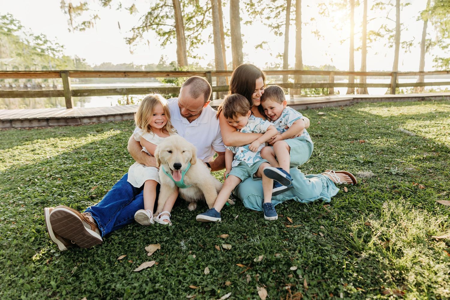 A family sits together on green grass with their small dog at sunset in a park with wooden fencing.