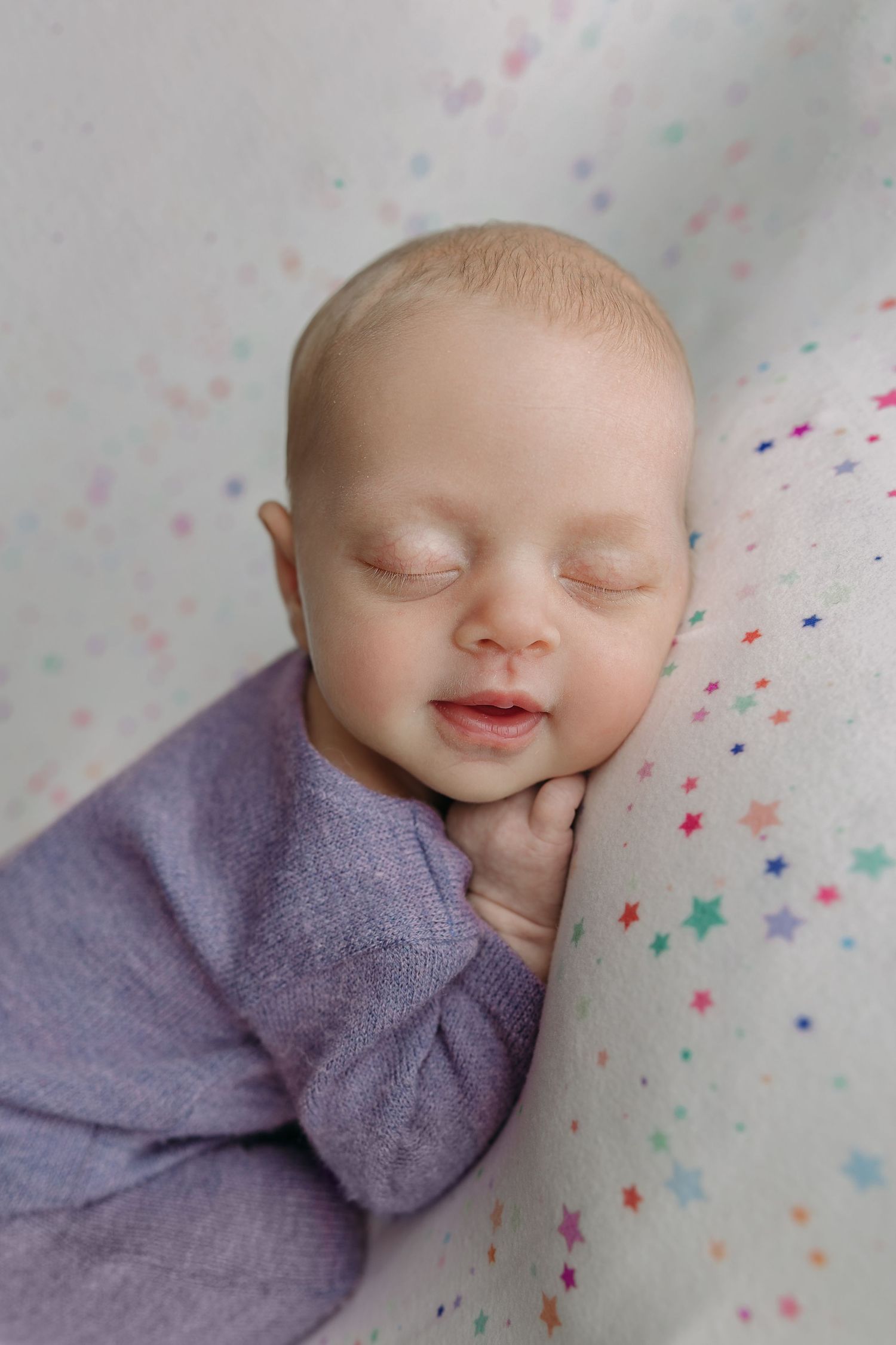 Sleeping newborn wearing purple wrap lies on pastel star-patterned fabric during peaceful studio photography session.