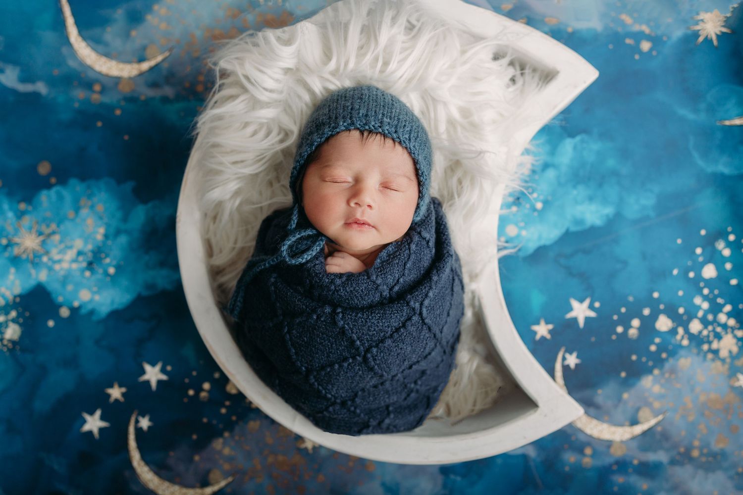 A sleeping baby wrapped in navy blue swaddle lies in a crescent moon prop against a starry blue background.