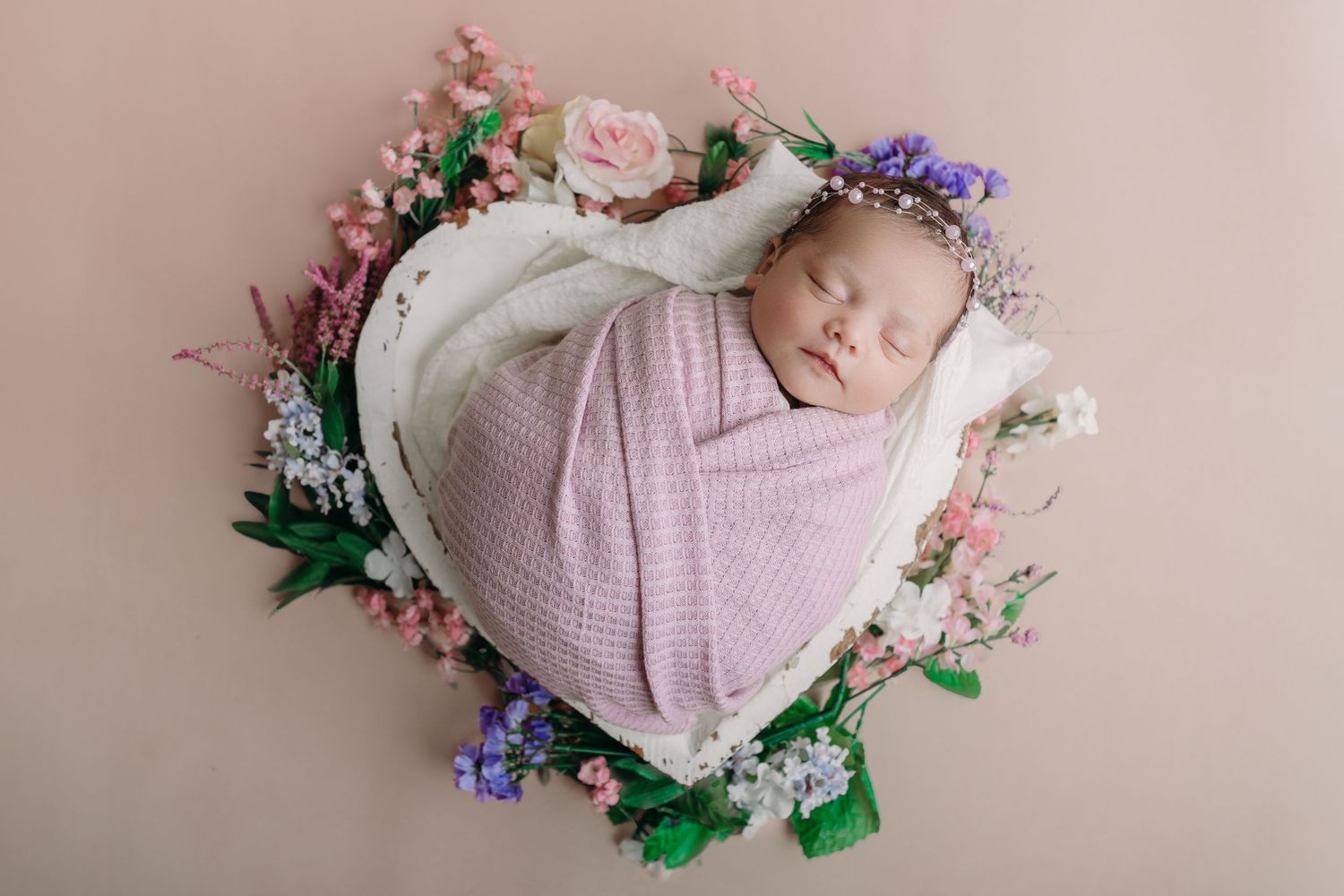 Newborn baby wrapped in soft lilac swaddle sleeping peacefully in circular floral wreath with pink and purple flowers.