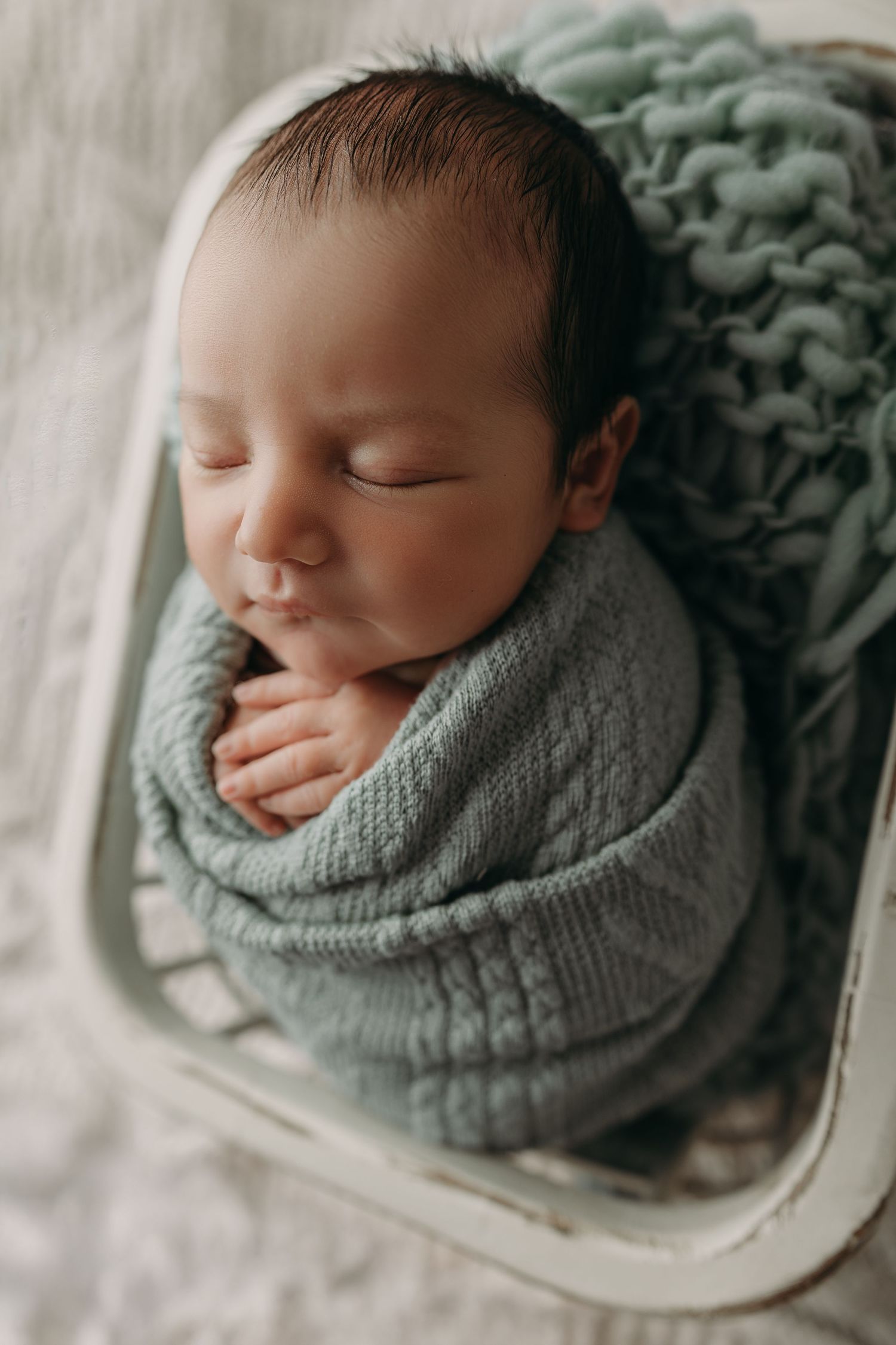 A sleeping newborn baby wrapped in a soft sage green knit blanket lies peacefully in a white bassinet.