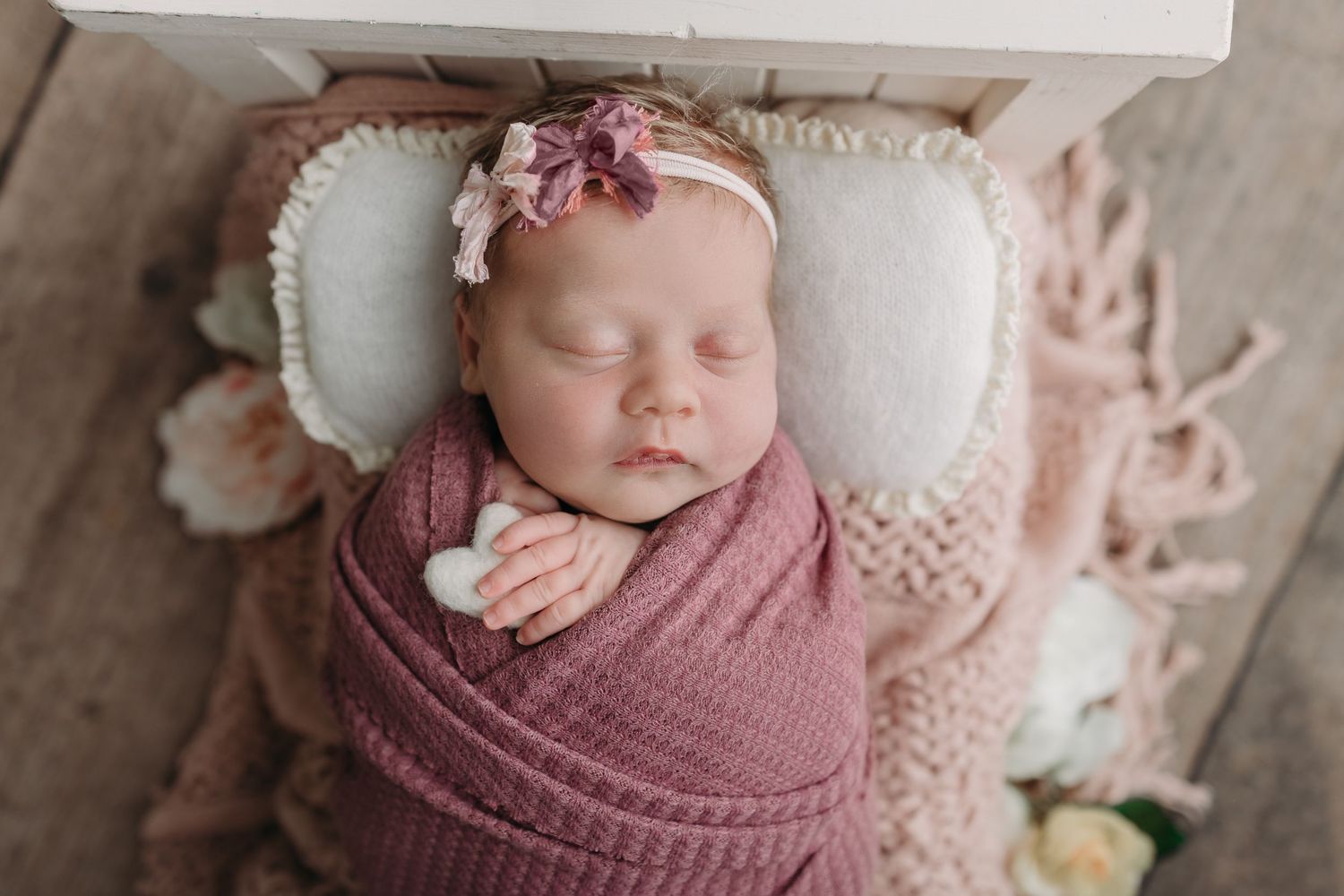 Sleeping newborn wrapped in mauve swaddle blanket resting on white pillow with pink floral headband.
