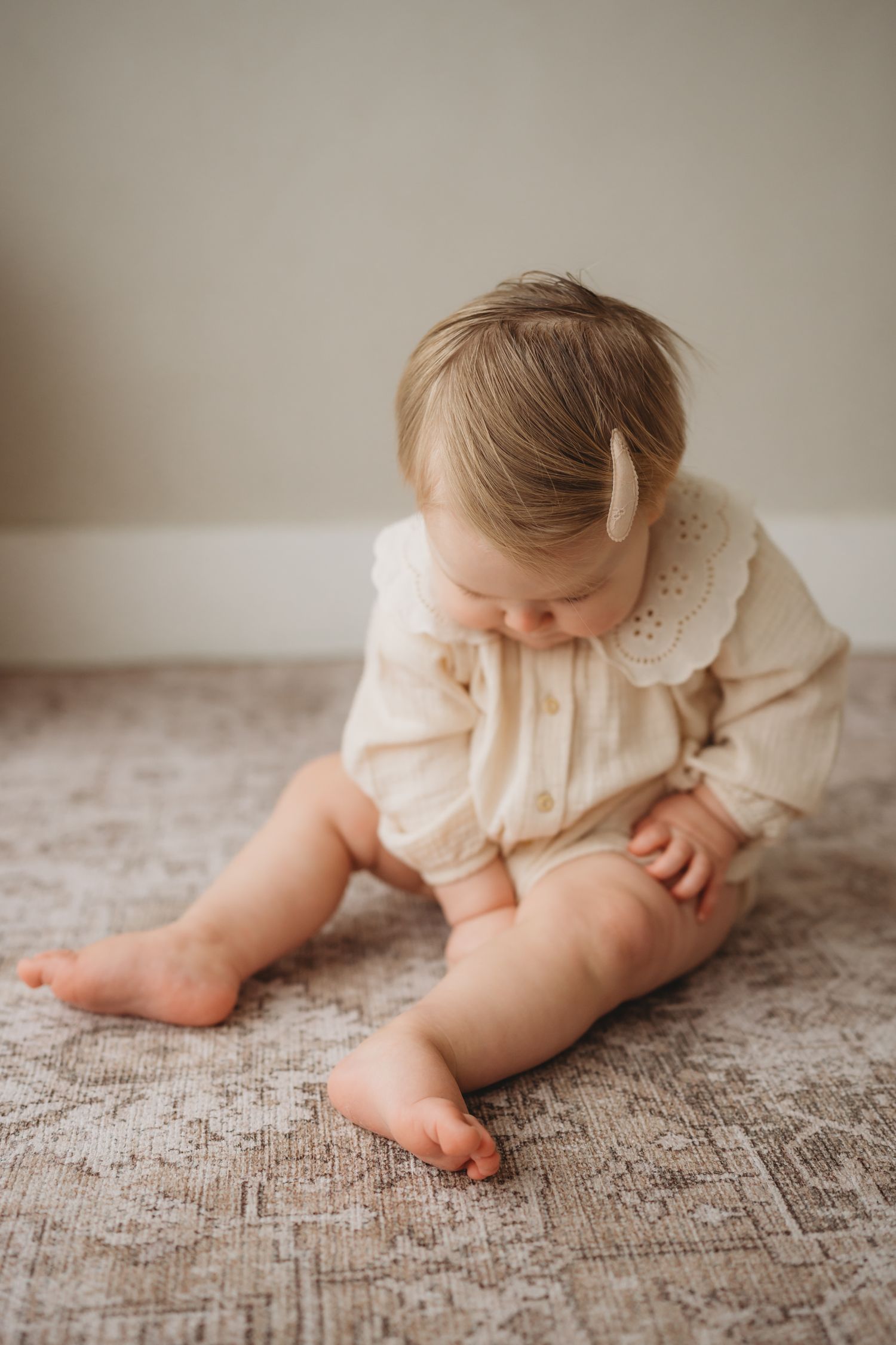 Baby in white dress sitting on textured carpet while looking down at their bare feet and toes.