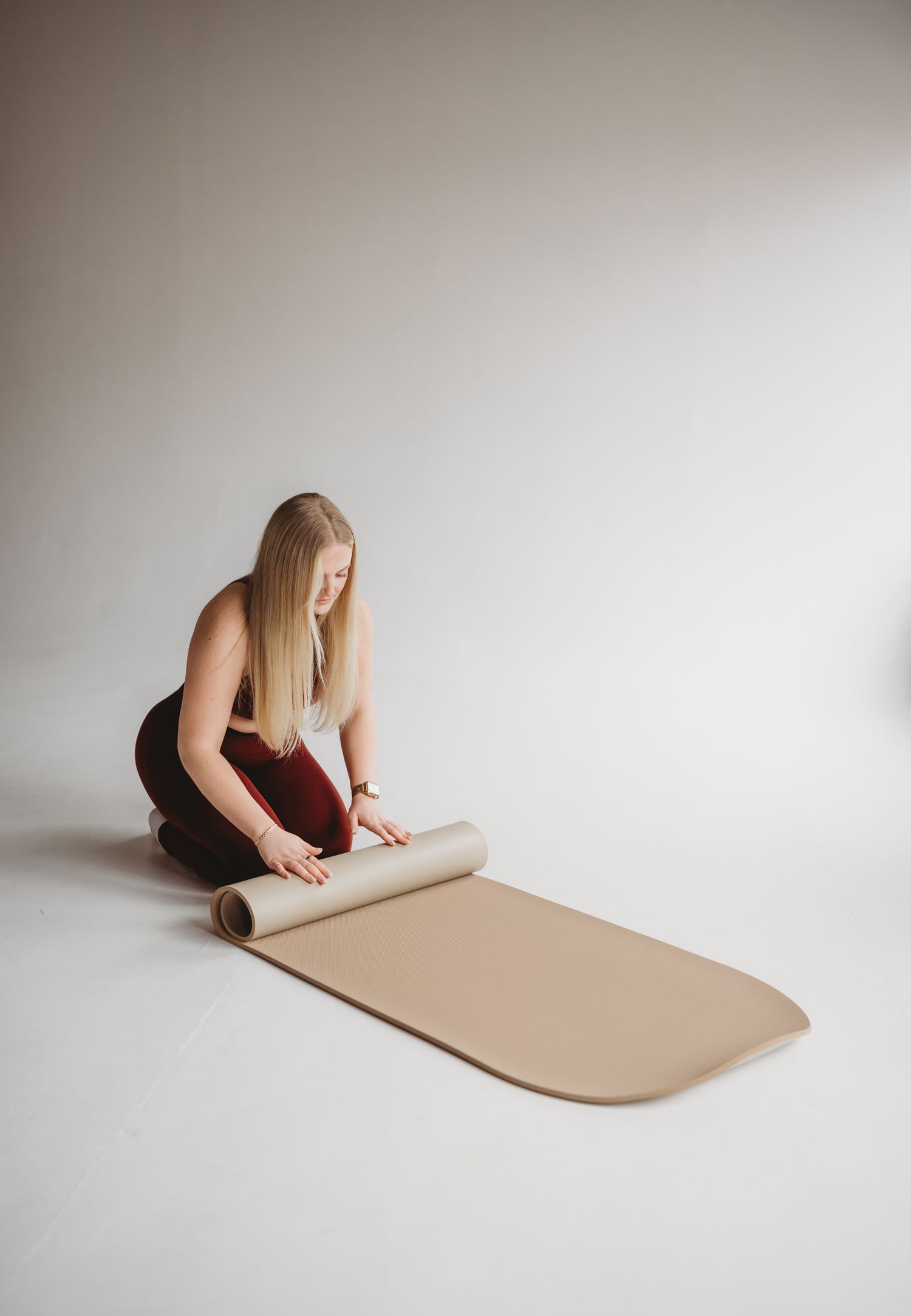 Person in black clothing unrolling a beige yoga or exercise mat on white studio floor.