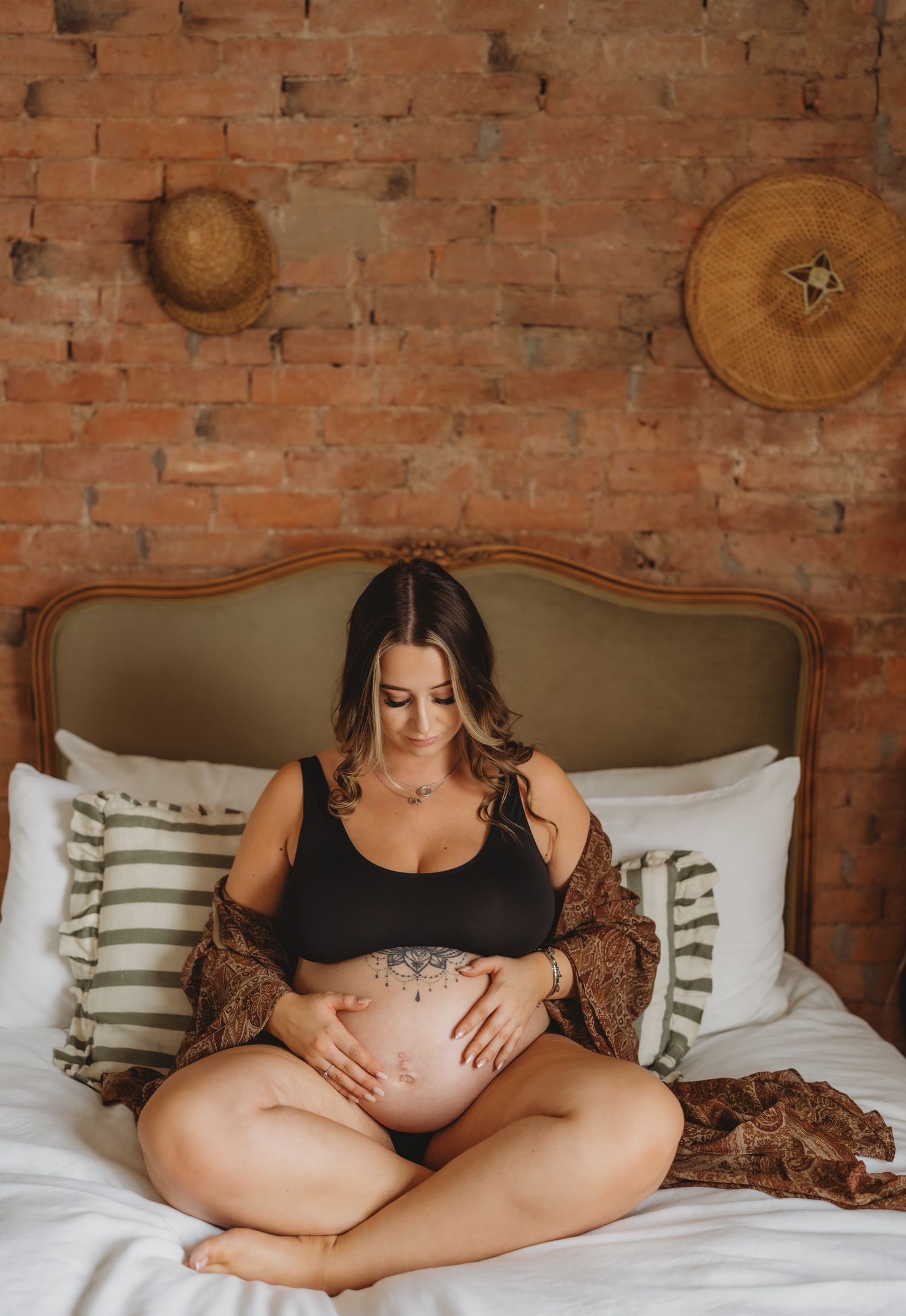 A maternity photo series showing a pregnant woman in black lounging on a bed against an exposed brick wall.