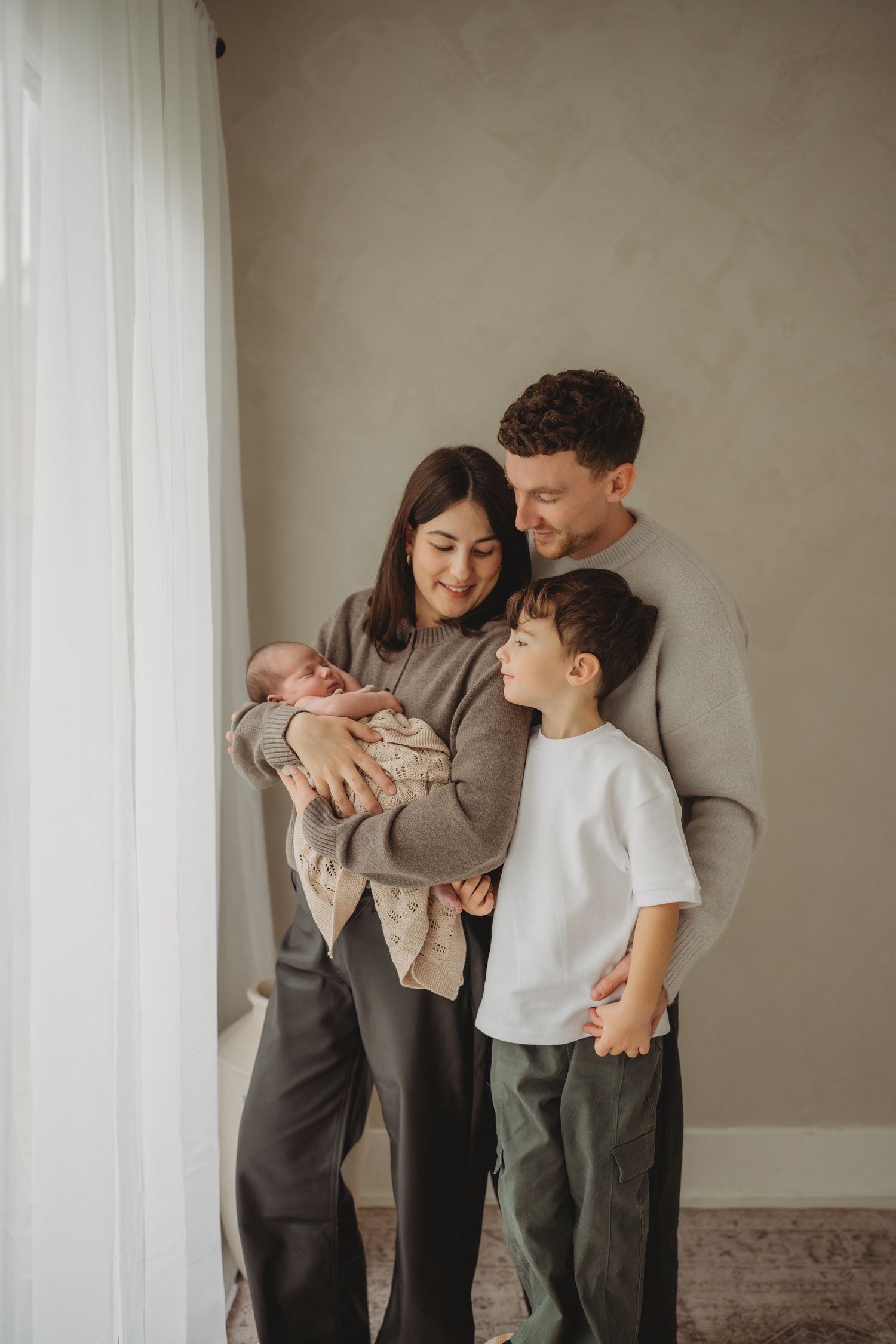 Family of four in neutral tones sharing tender moments against a minimalist white wall background.