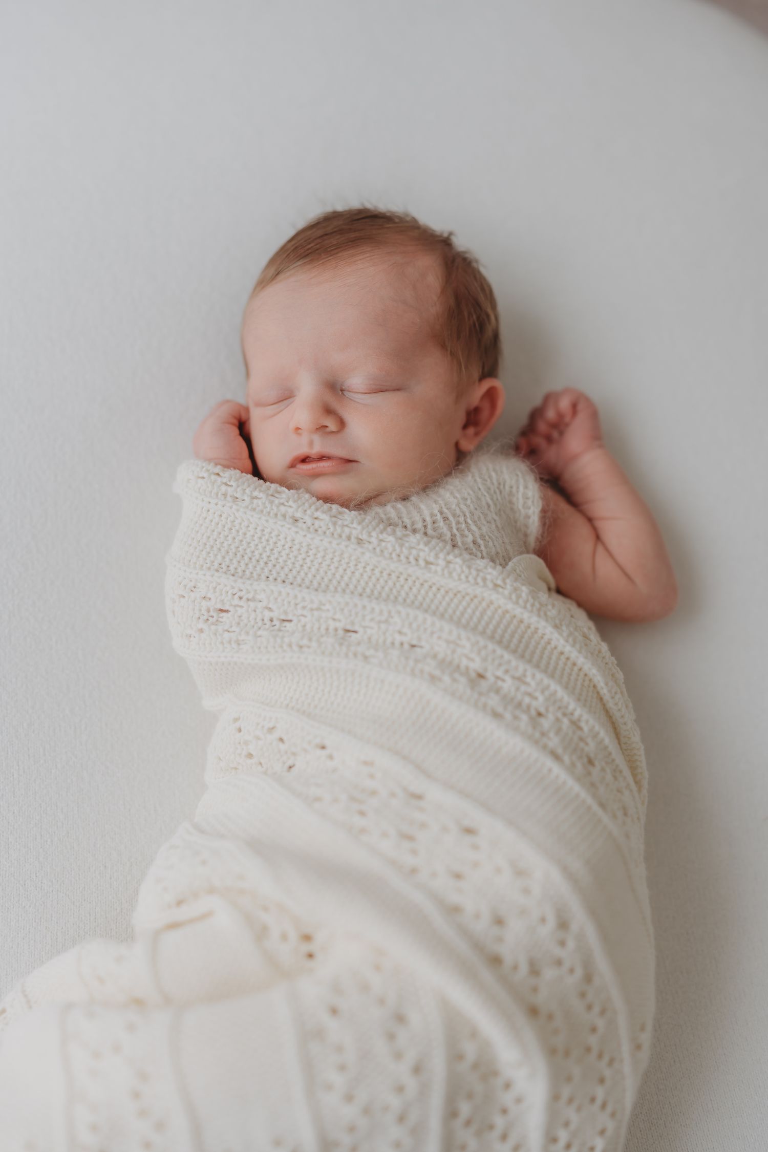 A sleeping newborn wrapped in a white lace blanket rests peacefully on a soft surface, during a newborn photoshoot in Leicestershire.