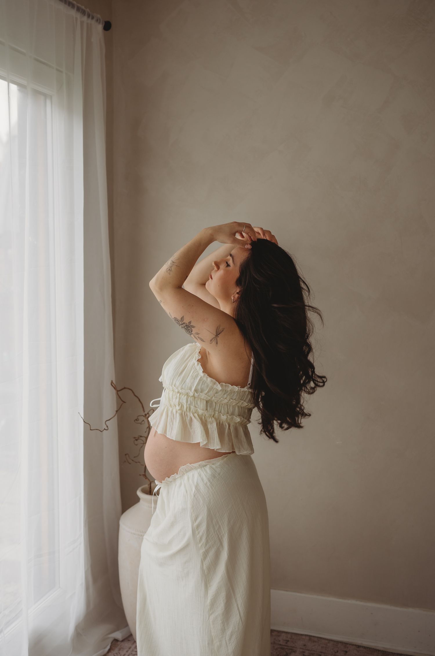 A pregnant woman in a white two-piece outfit stands near a window with soft natural lighting creating a dreamy atmosphere.