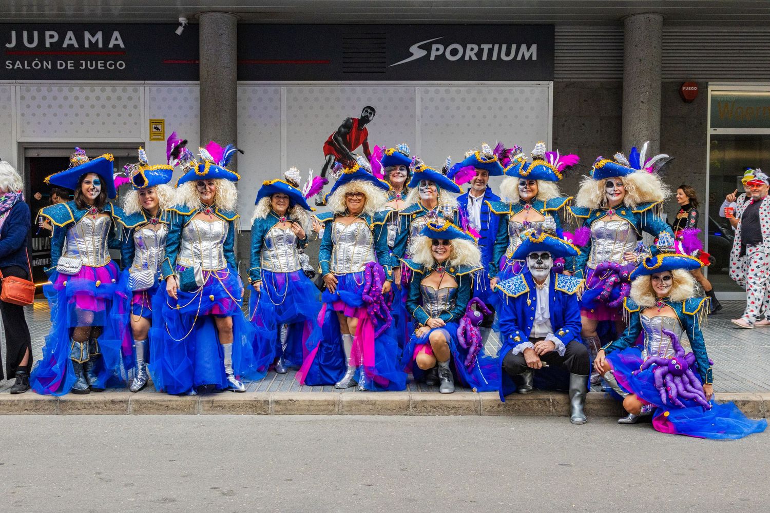 A group of dancers in blue and pink costumes perform a synchronized dance routine in a covered outdoor space.