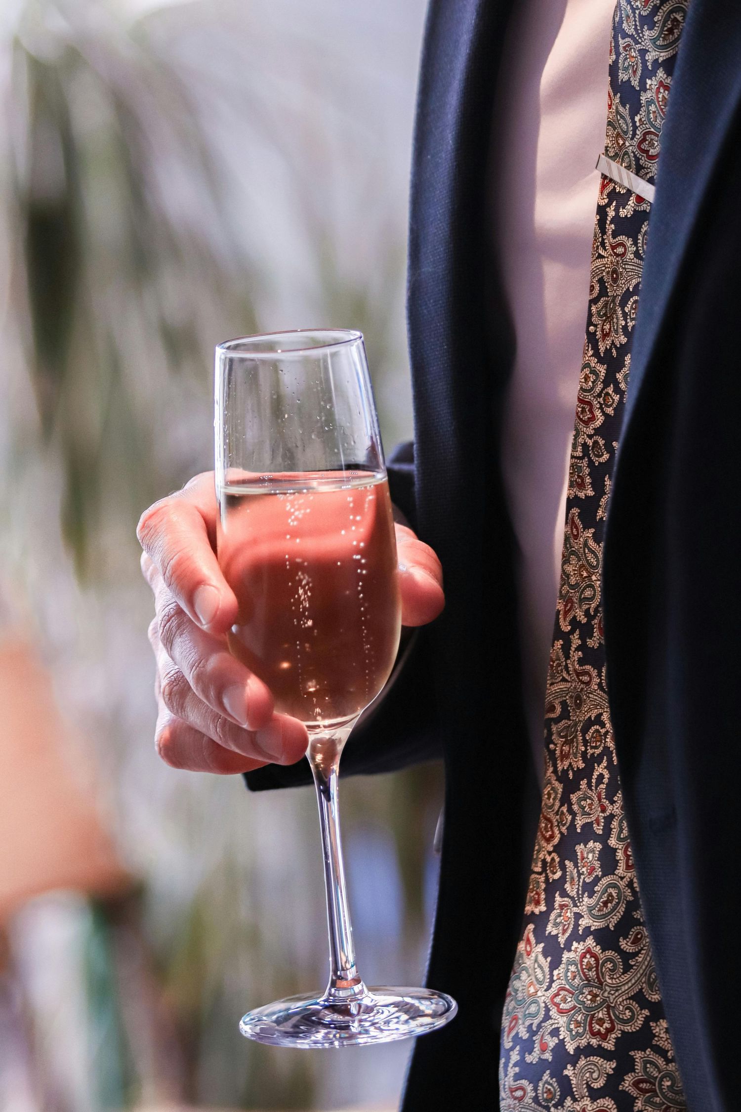 Hand in formal attire holds a delicate wine glass filled with pink rosé against a dark background.
