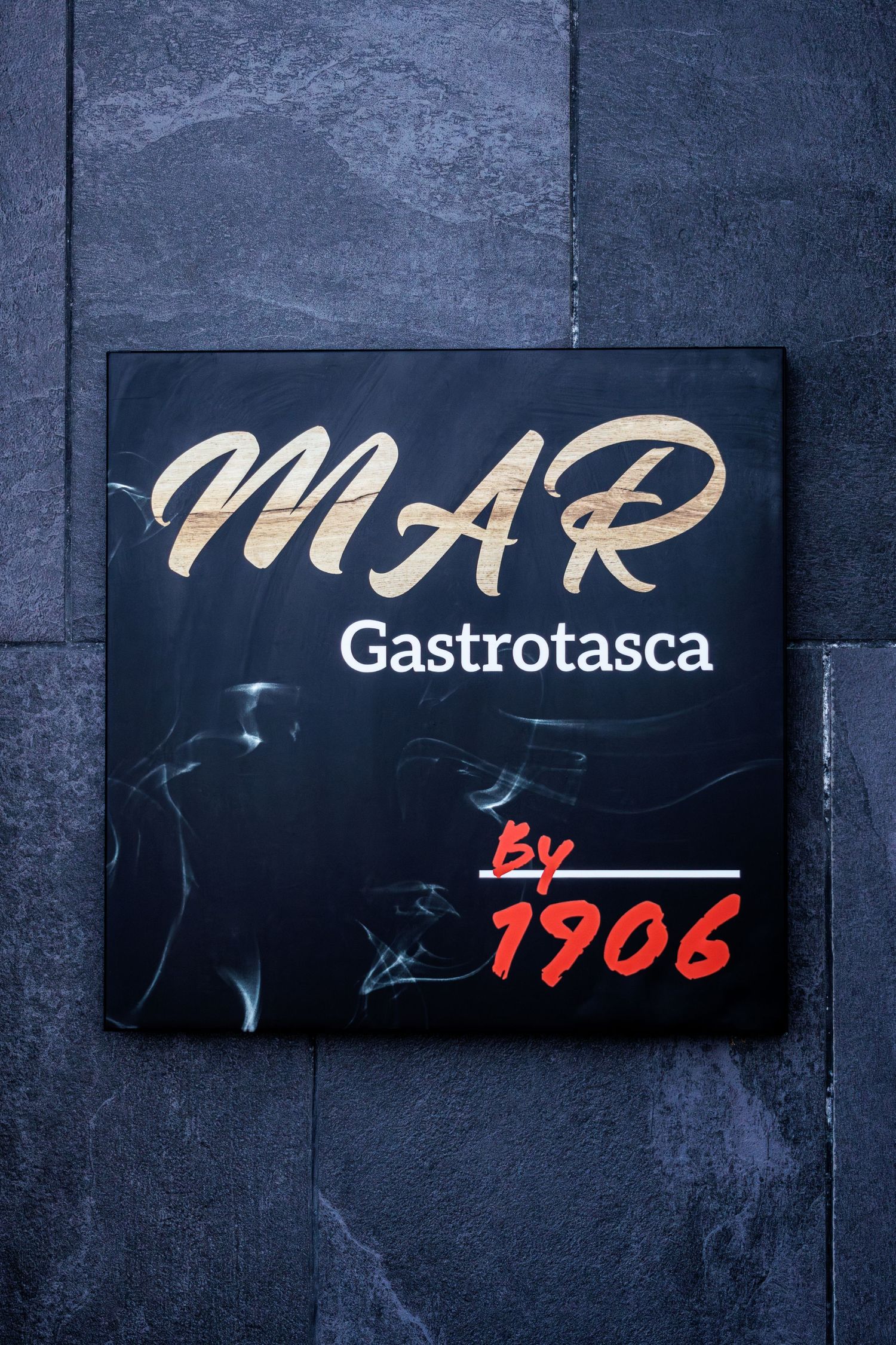 Black sign for MAR Gastrotasca by 1906 restaurant mounted on a dark slate stone wall.