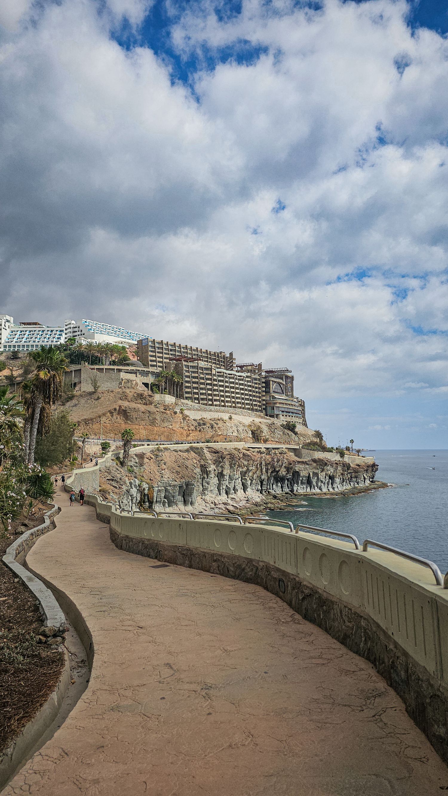 Curved coastal walkway along rocky cliffs with hotels and buildings overlooking the ocean in Gran Canaria.