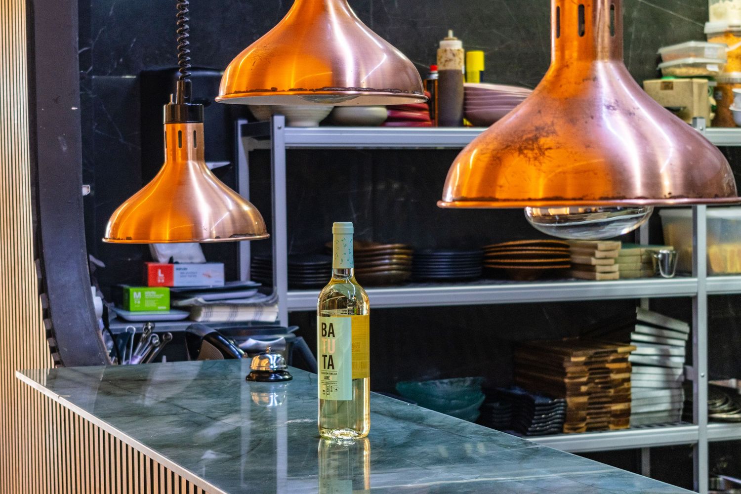 Copper pendant lights hang over a marble bar counter with a green bottle, shelves stocked with plates and condiments behind.